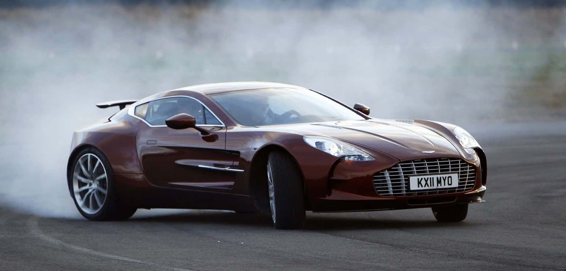 Aston Martin One-77 桌布 壁紙 — 一架時尚的棕色 Aston Martin 跑車在賽道上飄移，輪胎冒出滾滾濃煙。