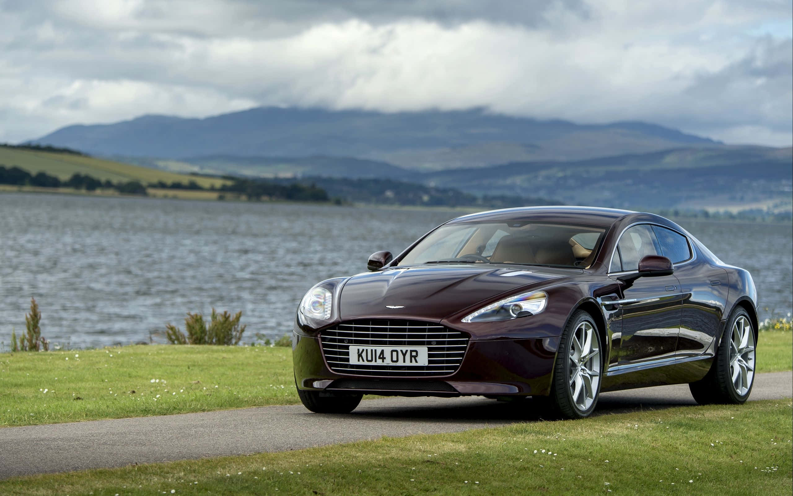 Aston Martin Rapide S 桌布 壁紙 — 一架時尚的棗紅色Aston Martin跑車停泊在湖邊的草地上，背景是起伏的山丘和多雲的天空。