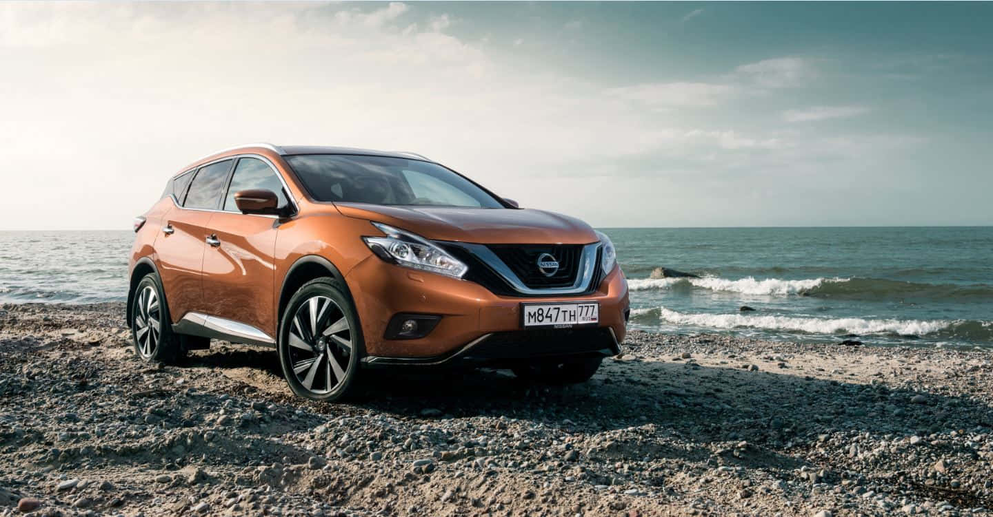 Nissan Murano 桌布 壁紙 — 一架時尚的橙色SUV停泊在岩石海灘上，海浪在附近拍打，天空多雲。