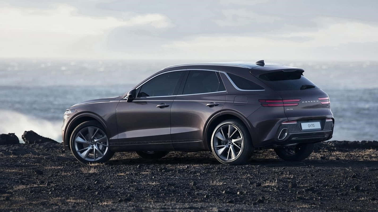 Genesis Gv70 桌布 壁紙 — 一架時尚的深灰色 Genesis GV70 SUV 停泊在黑色火山岩上，旁邊是波濤洶湧的海洋，天空多雲。