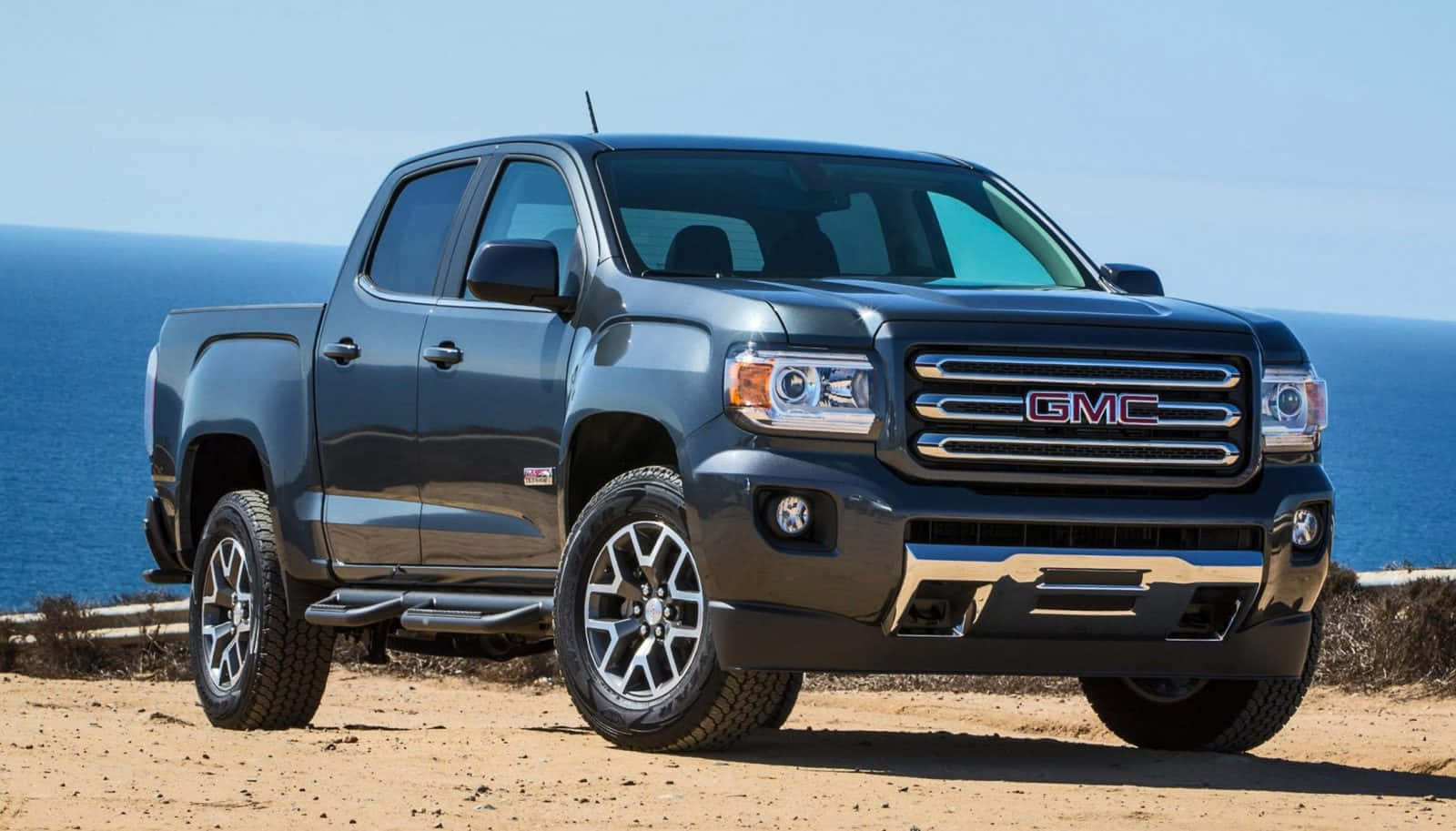 Gmc Canyon 桌布 壁紙 — 一架時尚的深灰色 GMC Canyon 皮卡車停泊在沙質懸崖上，俯瞰著晴朗天空下廣闊的藍色海洋。