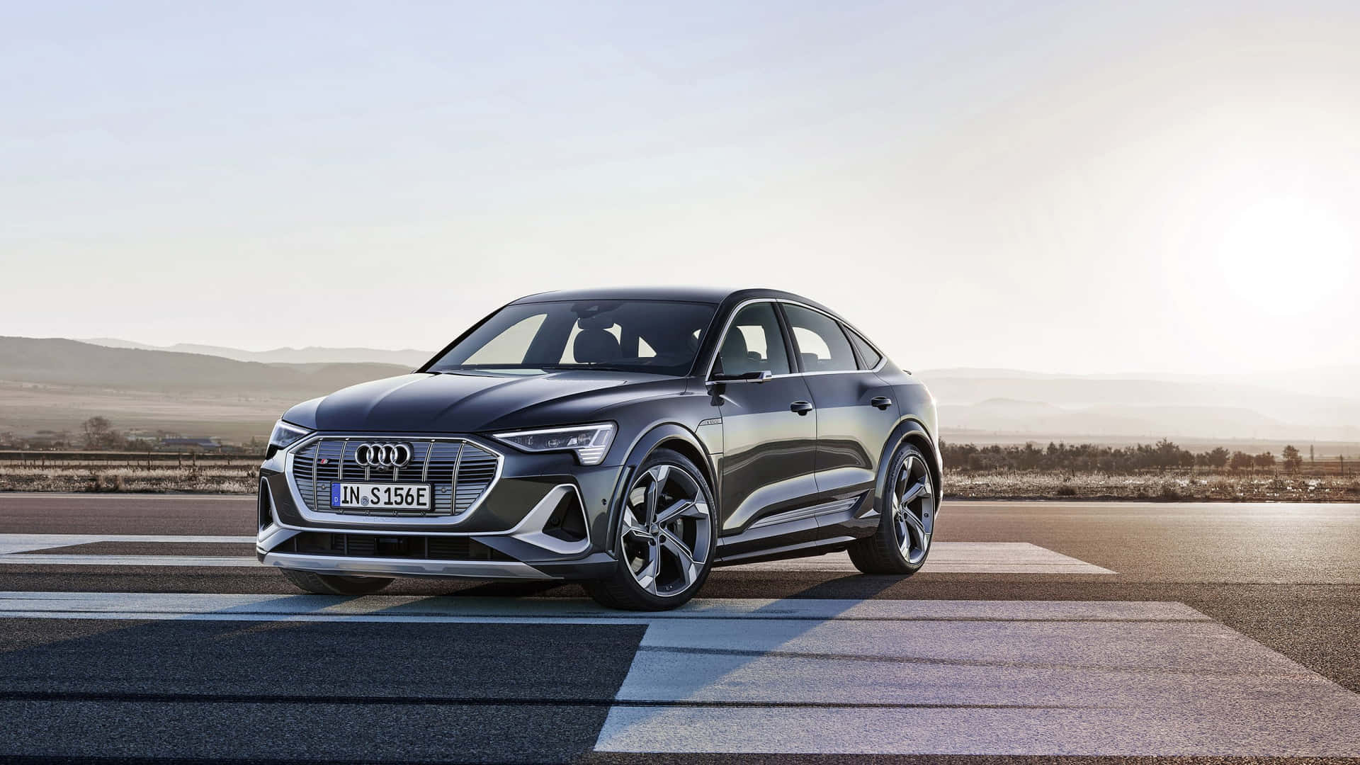  壁紙 — 一架時尚的深灰色Audi e-tron S停在陽光普照的道路上，背景是連綿起伏的山丘。