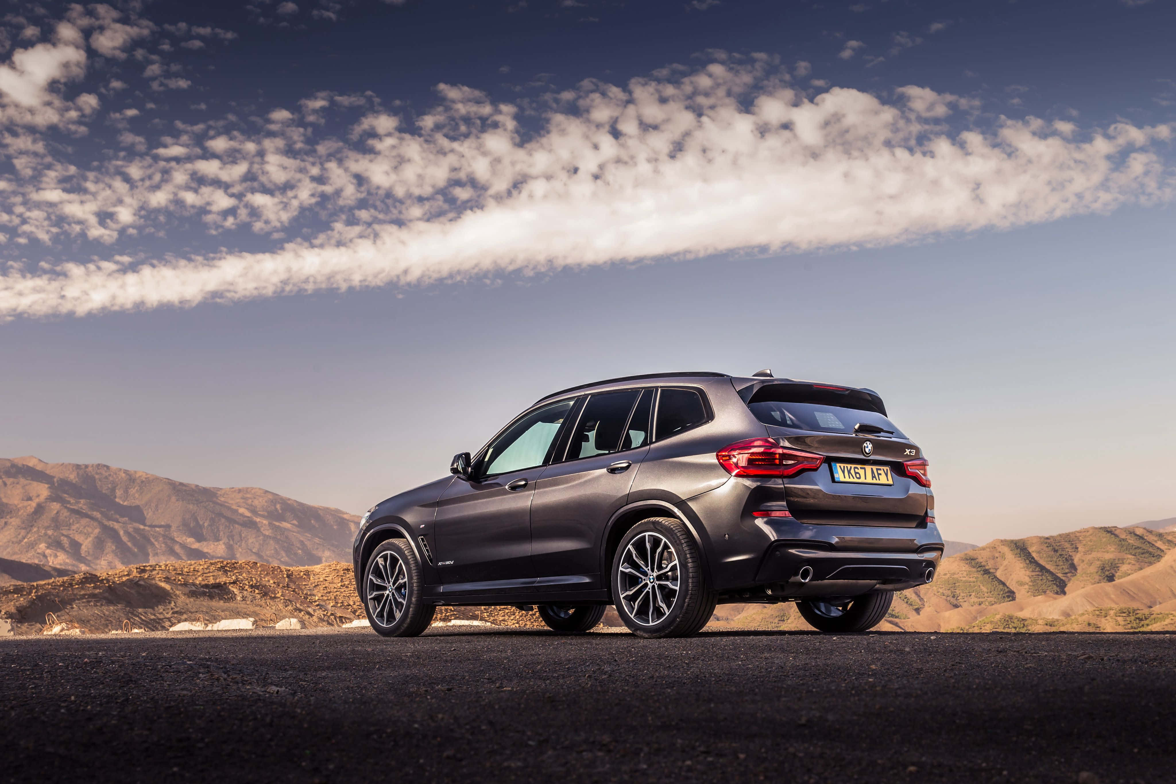 Bmw X3 桌布 壁紙 — 一架時尚的深灰色BMW X3 SUV停泊在沙漠公路上，背景是起伏的山丘和部分多雲的天空。