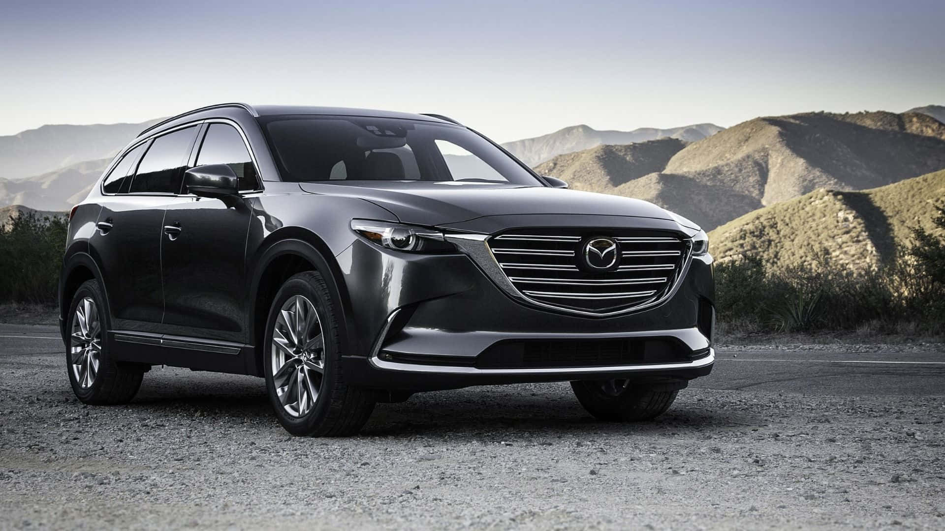 Mazda Cx-9 桌布 壁紙 — 一架時尚的深灰色Mazda CX-9 SUV停泊在碎石路上，背景是乾旱的山丘和朦朧的天空。