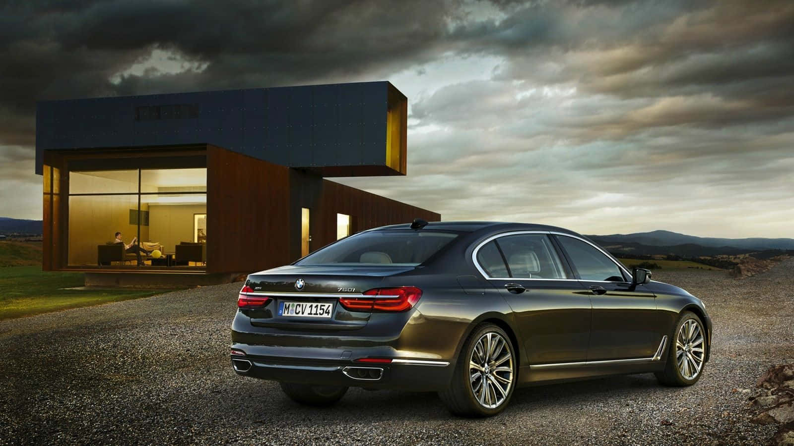 Bmw 7 Series 桌布 壁紙 — 一架時尚的深灰色寶馬7系轎車停泊在碎石路上，旁邊是一座現代木屋，背景是戲劇性的多雲天空。
