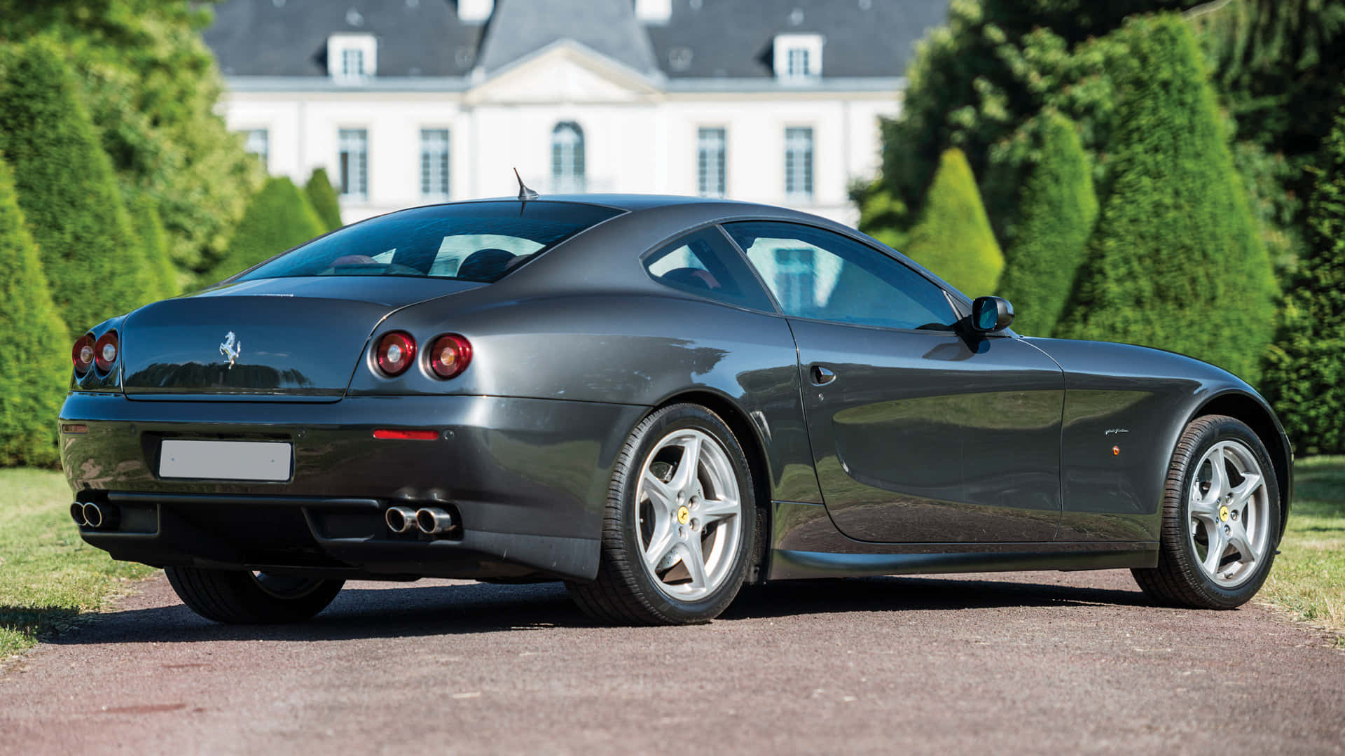 Ferrari 612 Scaglietti 桌布 壁紙 — 一架時尚的深灰色法拉利跑車停泊在碎石車道上，背景是一座宏偉的白色豪宅和修剪整齊的綠色樹籬。