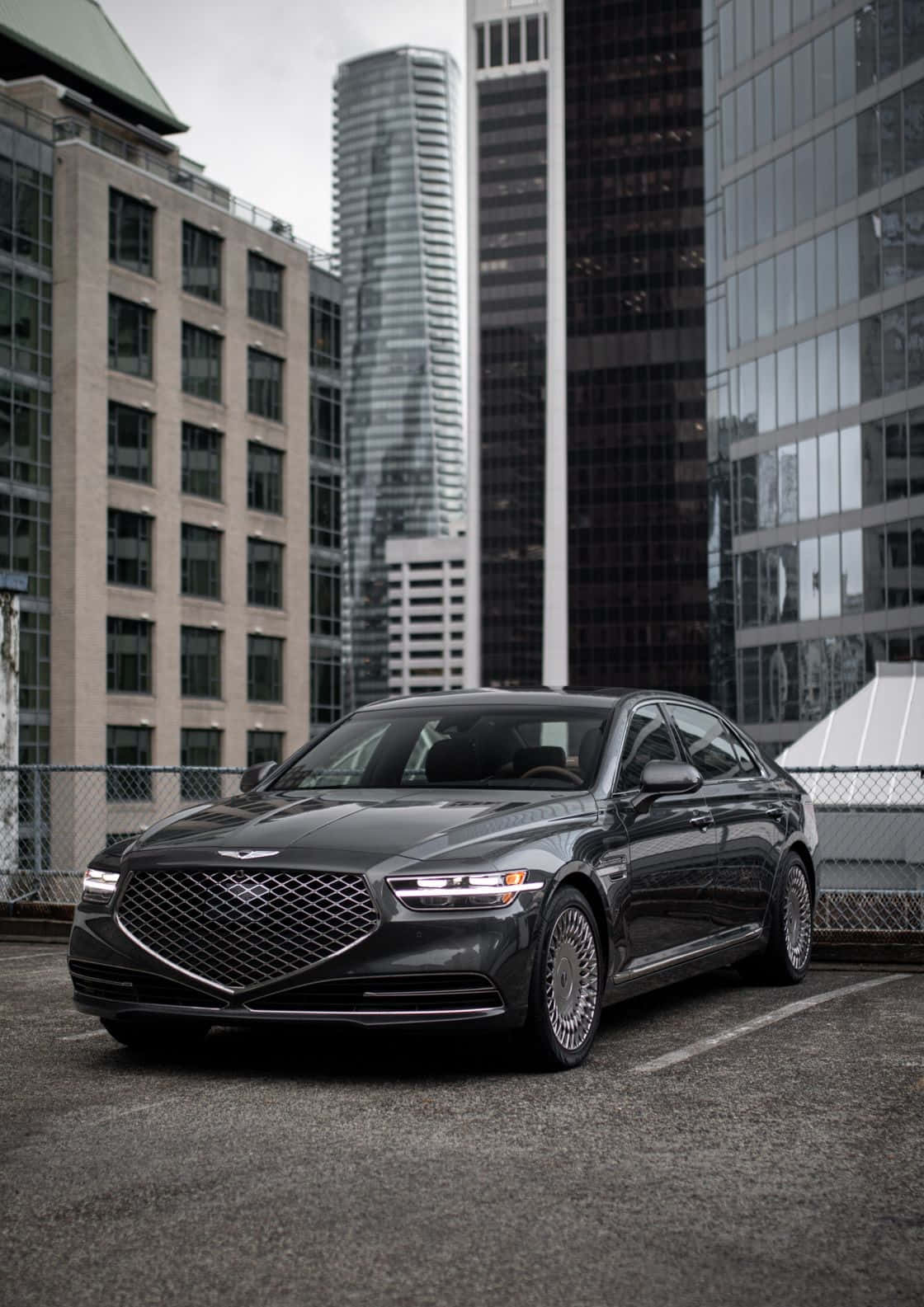 Genesis G90 桌布 壁紙 — 一架時尚的深灰色豪華轎車停泊在屋頂上，俯瞰著現代城市天際線，高聳的玻璃和混凝土建築林立。