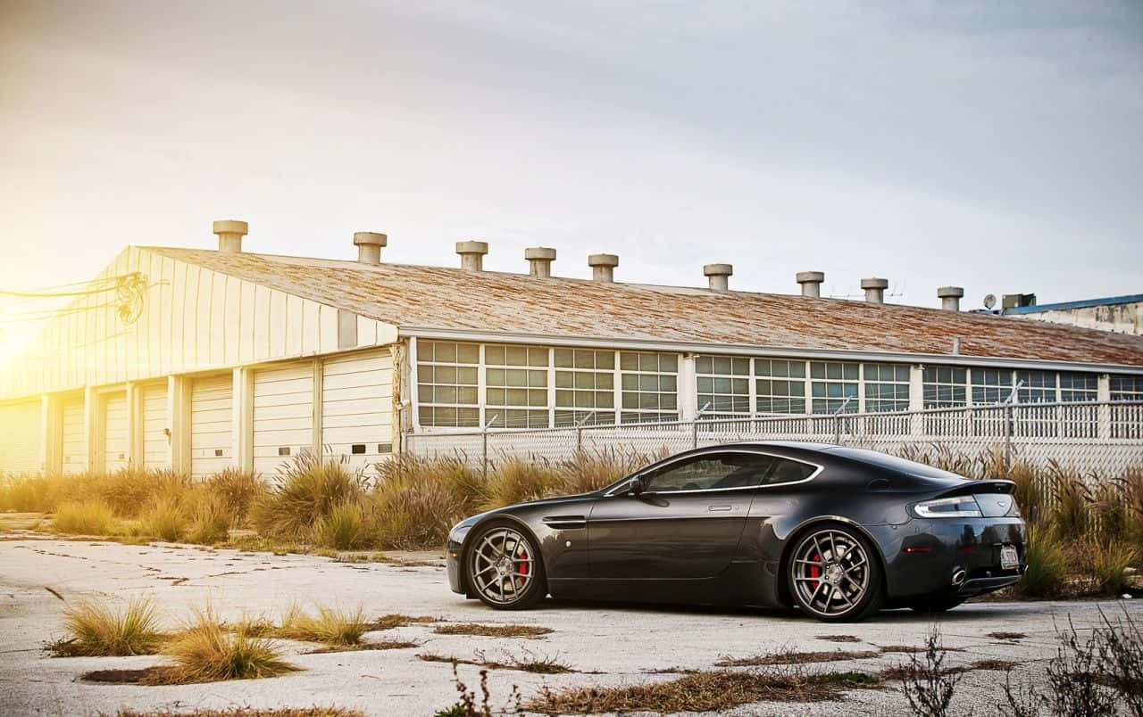 Aston Martin V12 Vantage 桌布 壁紙 — 一架時尚的深灰色跑車在黃金時刻停泊在風化的工業建築前。