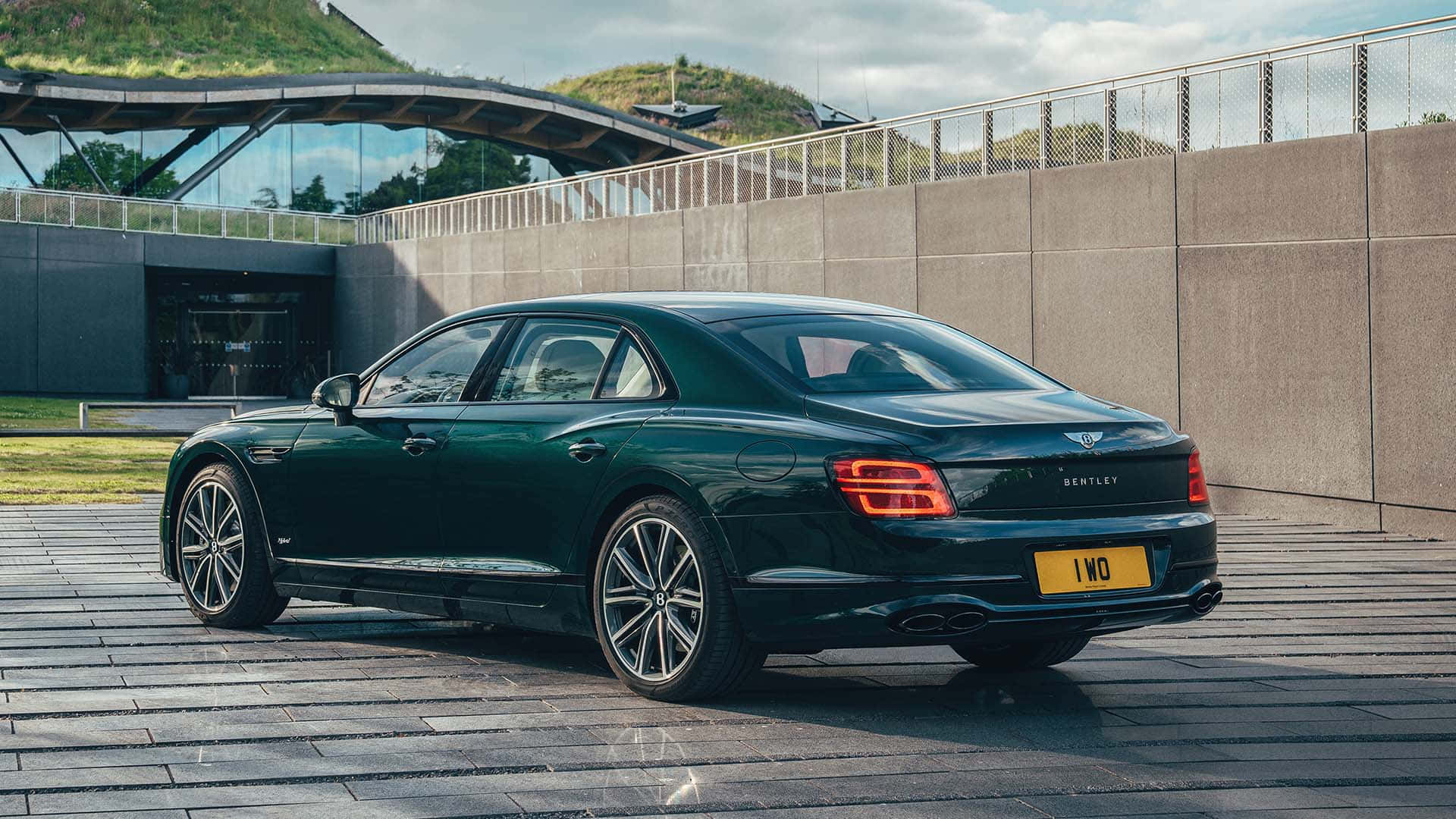 Bentley Flying Spur 桌布 壁紙 — 一架時尚的深綠色賓利飛馳停在石頭廣場上，旁邊是現代玻璃建築，背景有草地山丘。