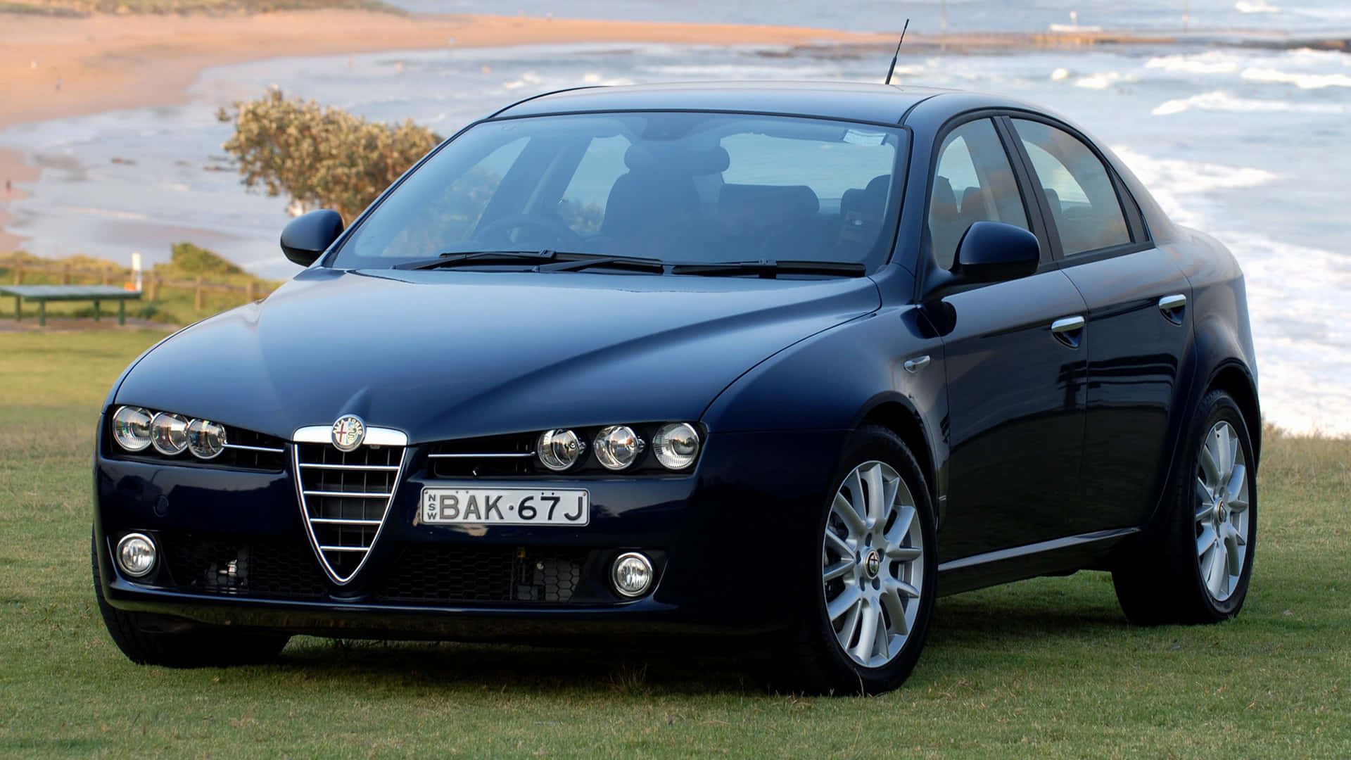 Alfa Romeo 159 桌布 壁紙 — 一架時尚的深藍色 Alfa Romeo 房車停泊在沙灘附近的草地上，背景有海浪拍打。