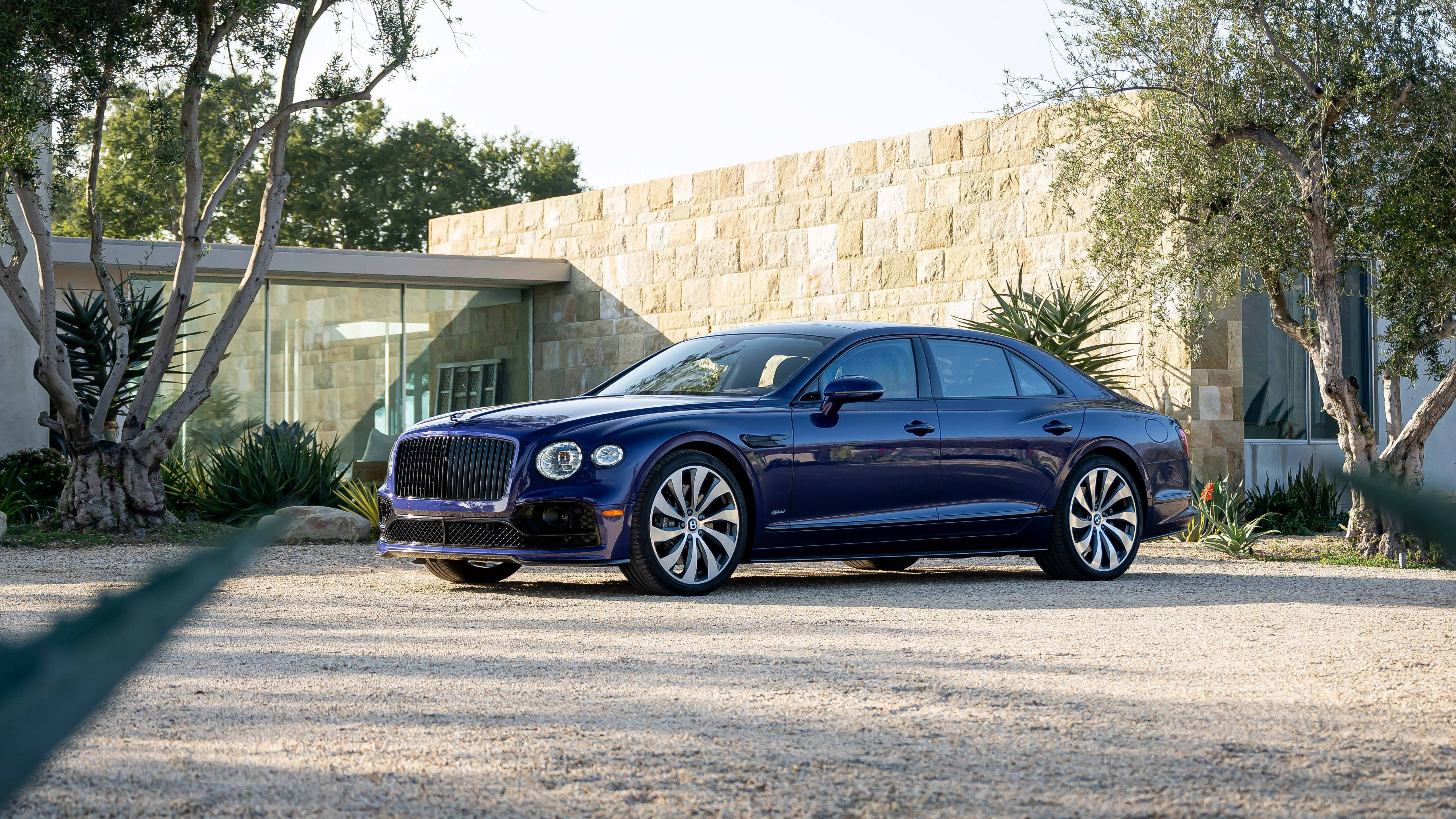 4K 桌布 壁紙 — 一架時尚的深藍色 Bentley Flying Spur 停泊在碎石車道上，前面是一座被樹木環繞的現代石頭和玻璃房屋。
