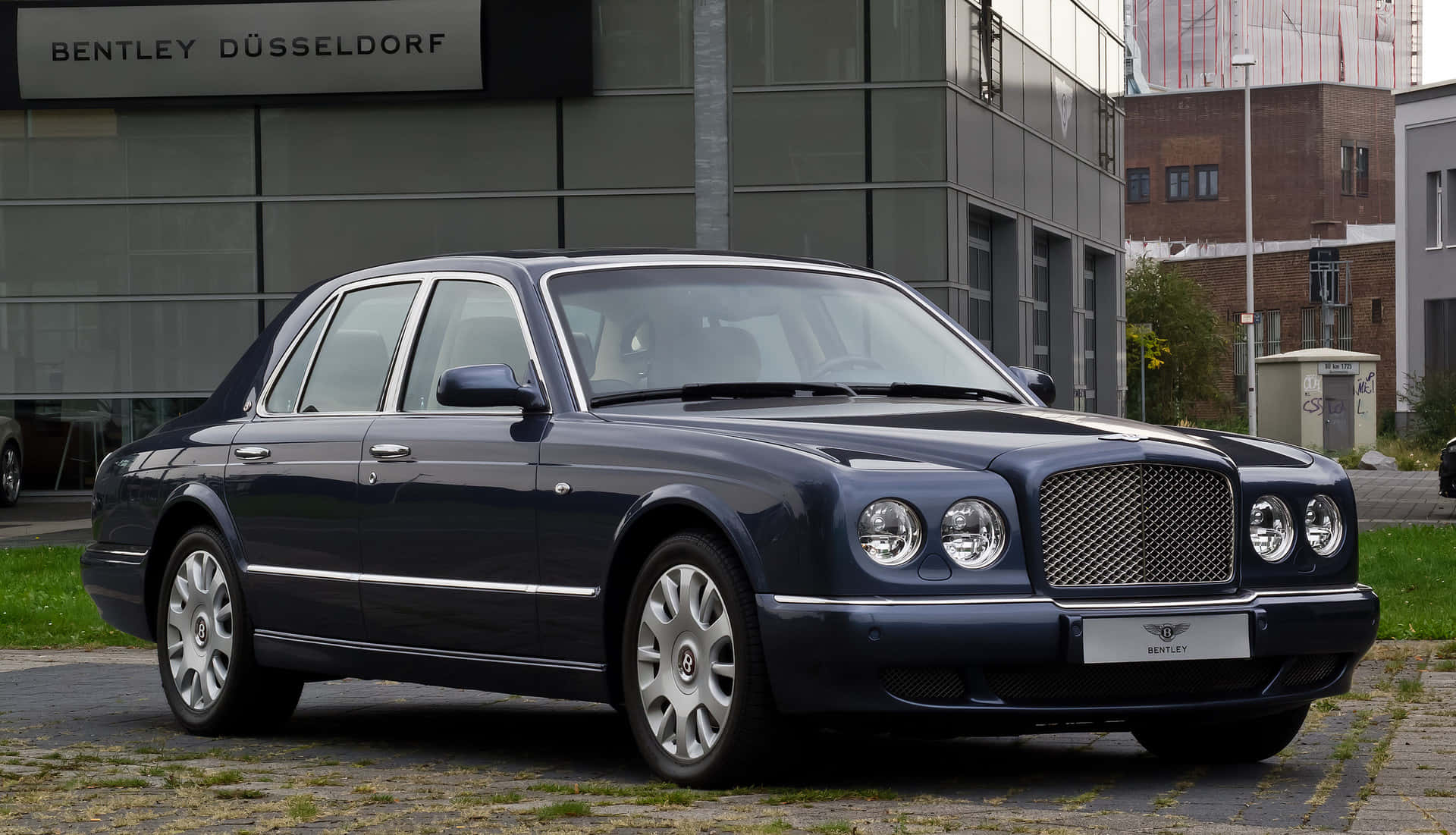 Bentley Arnage 桌布 壁紙 — 一架時尚的深藍色賓利房車停泊在鵝卵石路上，前面是現代玻璃建築，帶有'BENTLEY DÜSSELDORF'標誌。
