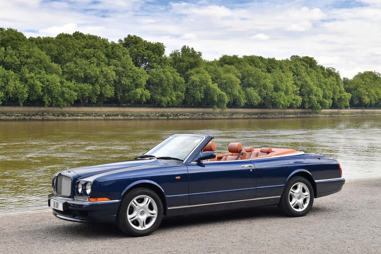 Bentley Azure 桌布 壁紙 — 一架時尚的深藍色賓利開篷車，配橙色皮革座椅，停泊在平靜的河邊，兩旁綠樹成蔭，天空局部多雲。