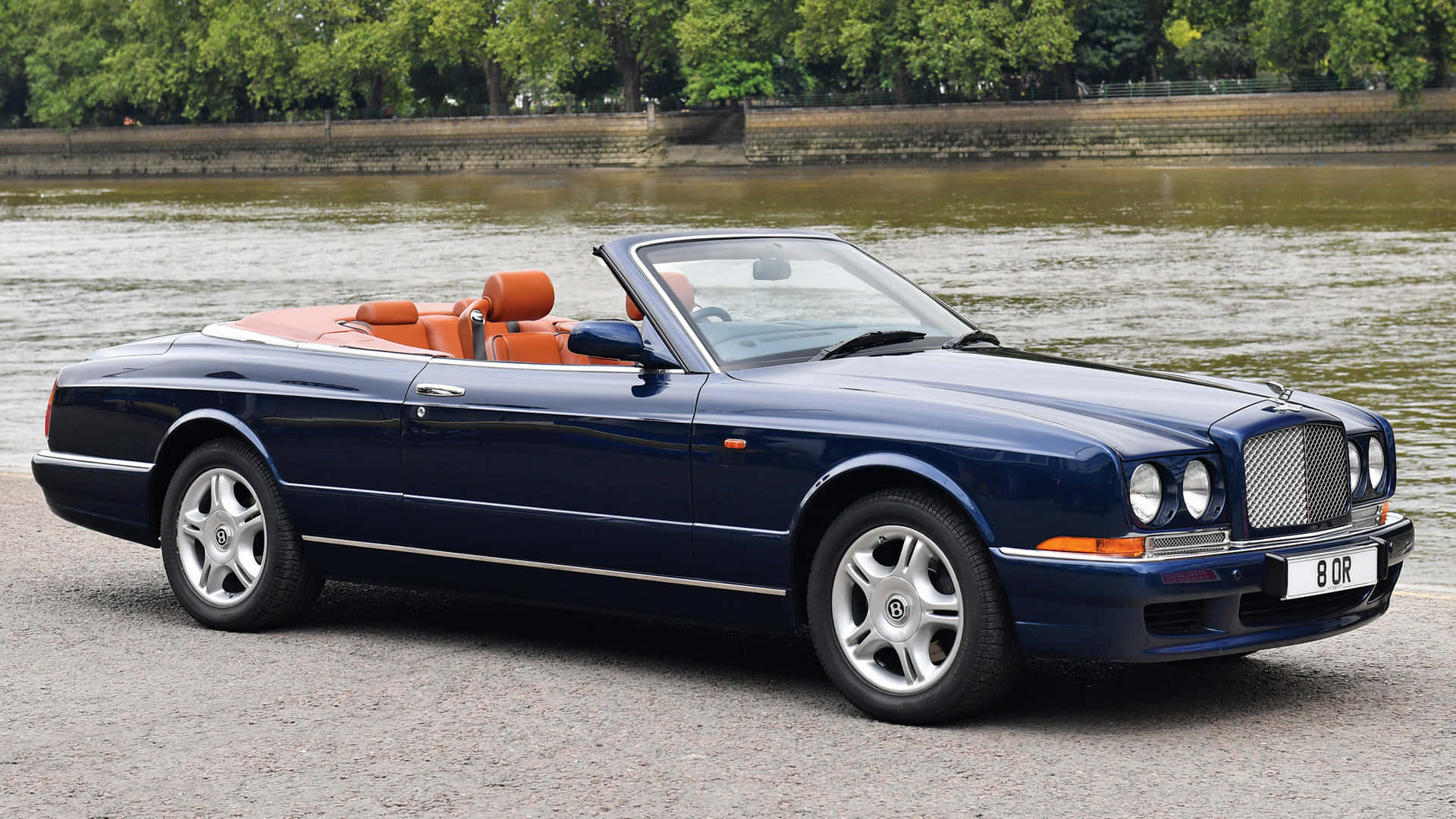 Bentley Azure 桌布 壁紙 — 一架時尚的深藍色賓利開篷車，配橙色皮革座椅，在日間停泊在河岸邊。