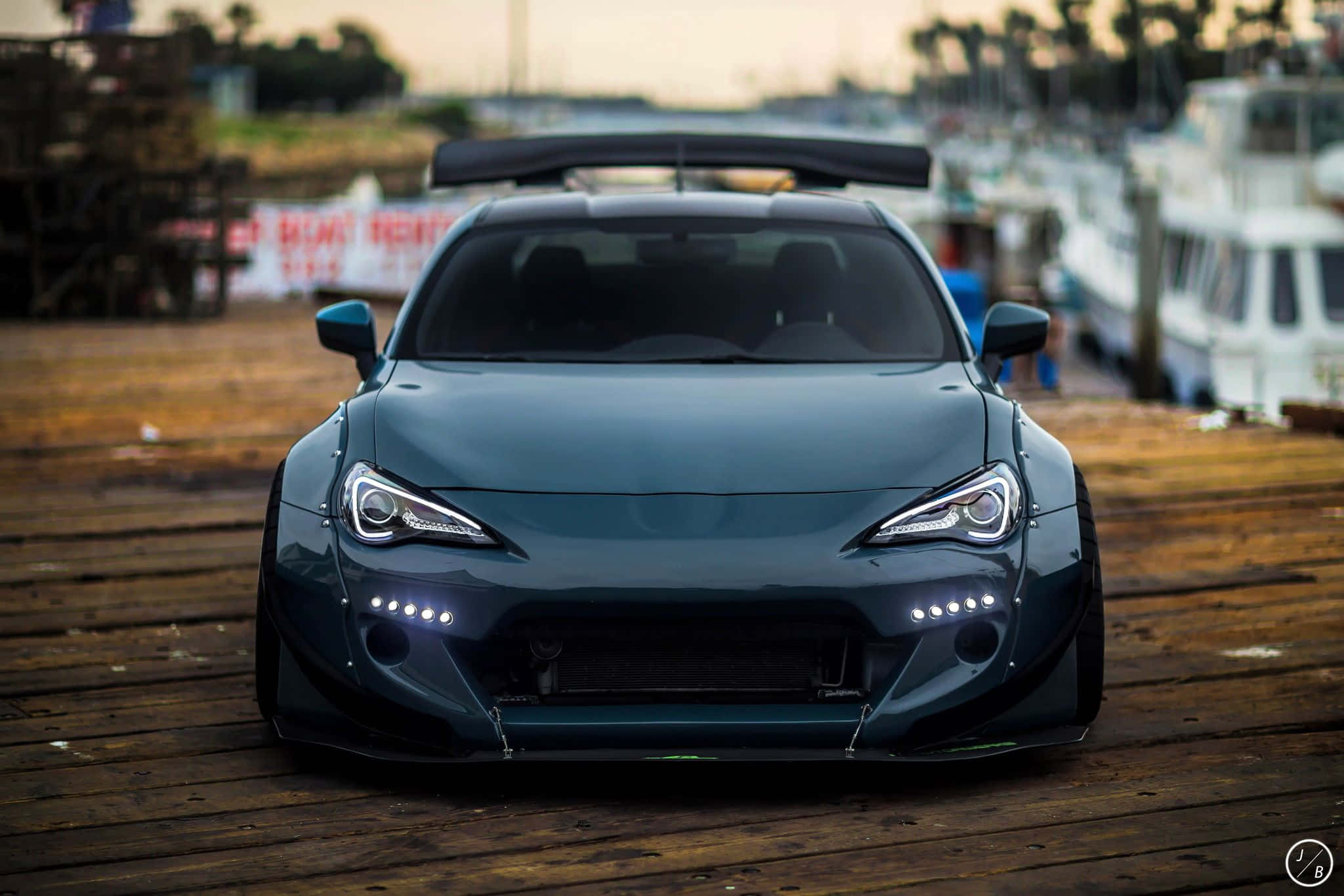 Toyota 86 壁紙 — 一架時尚的深青色改裝跑車，亮著車頭燈，停泊在黃昏時分風化的木製碼頭上，背景有船隻。