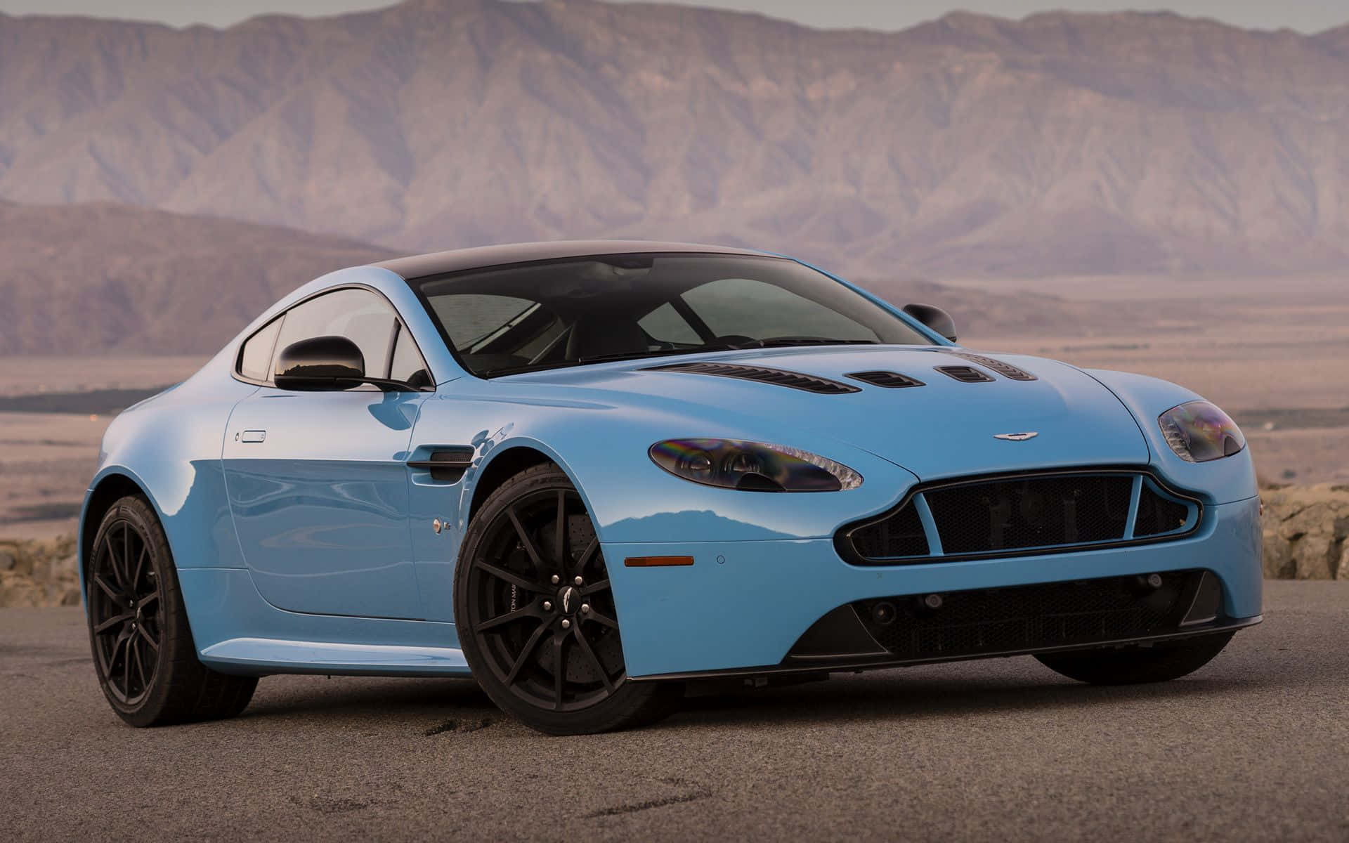 Aston Martin V12 Vantage 桌布 壁紙 — 一架時尚的淺藍色 Aston Martin 跑車停泊在沙漠公路上，背景是崎嶇的山脈。