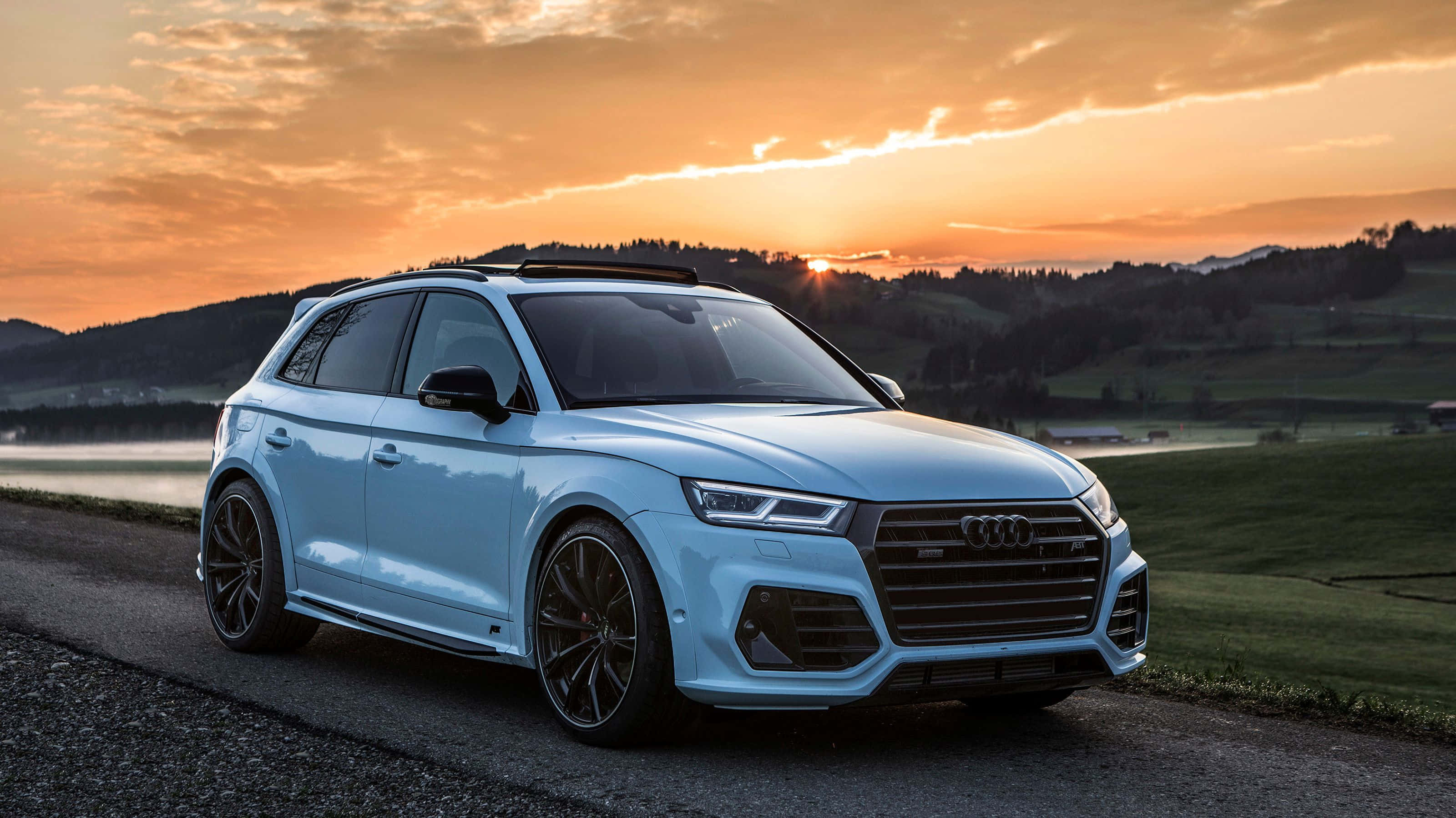Audi Sq5 桌布 壁紙 — 一架時尚的淺藍色 Audi SQ5 SUV 停泊在日落時分的鄉村道路上，背後是金色的天空和起伏的綠色山丘。