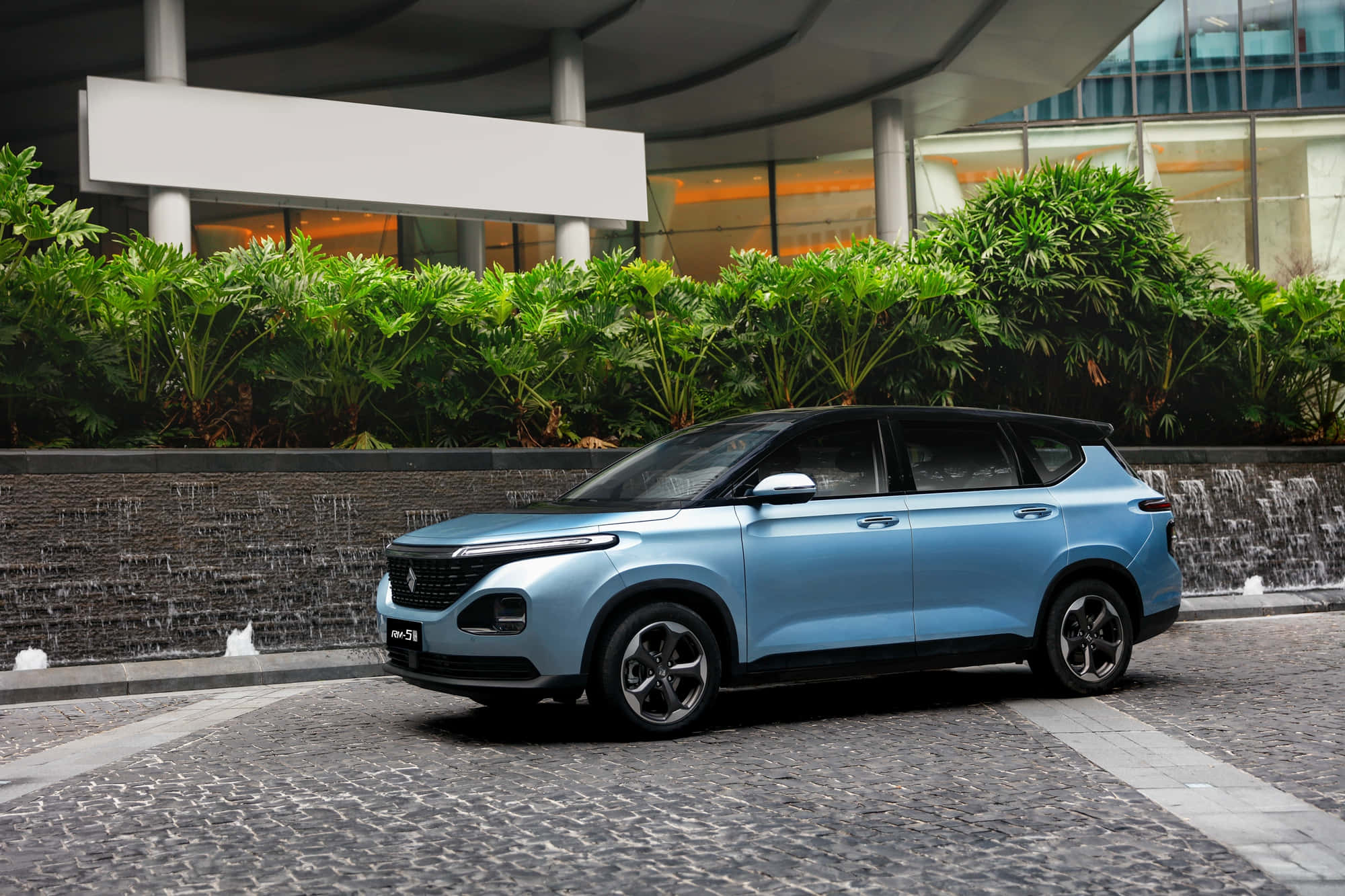 Baojun 桌布 壁紙 — 一架時尚的淺藍色SUV停泊在鵝卵石街道上，旁邊是茂密的綠植和一座帶有溫暖室內照明的現代建築。