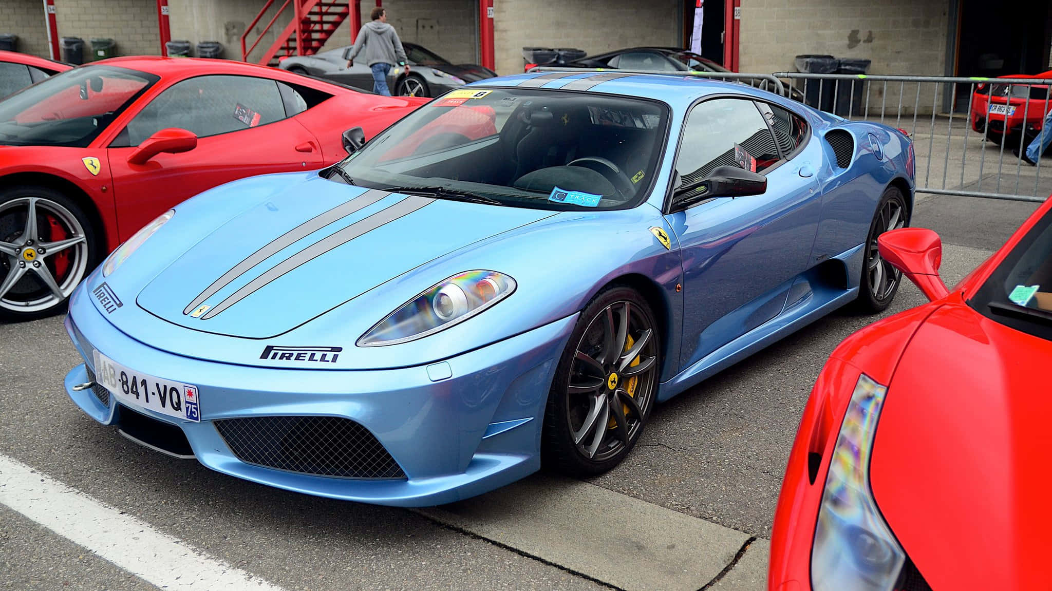 Ferrari F430 桌布 壁紙 — 一架時尚的淺藍色法拉利跑車，帶有灰色賽車條紋，停泊在車庫中的紅色法拉利之間。