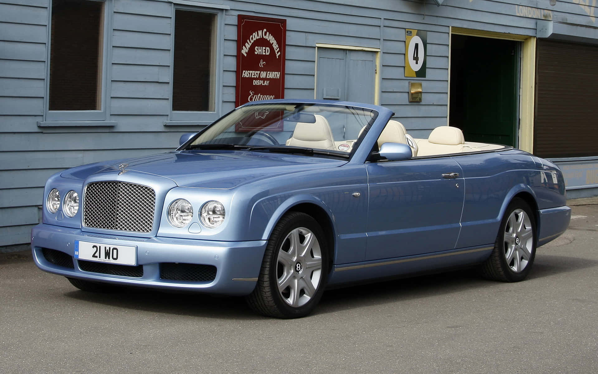 Bentley Azure 桌布 壁紙 — 一架時尚的淺藍色賓利開篷車，配以奶油色內飾，停在一個帶有復古賽車標誌的藍色木棚外。