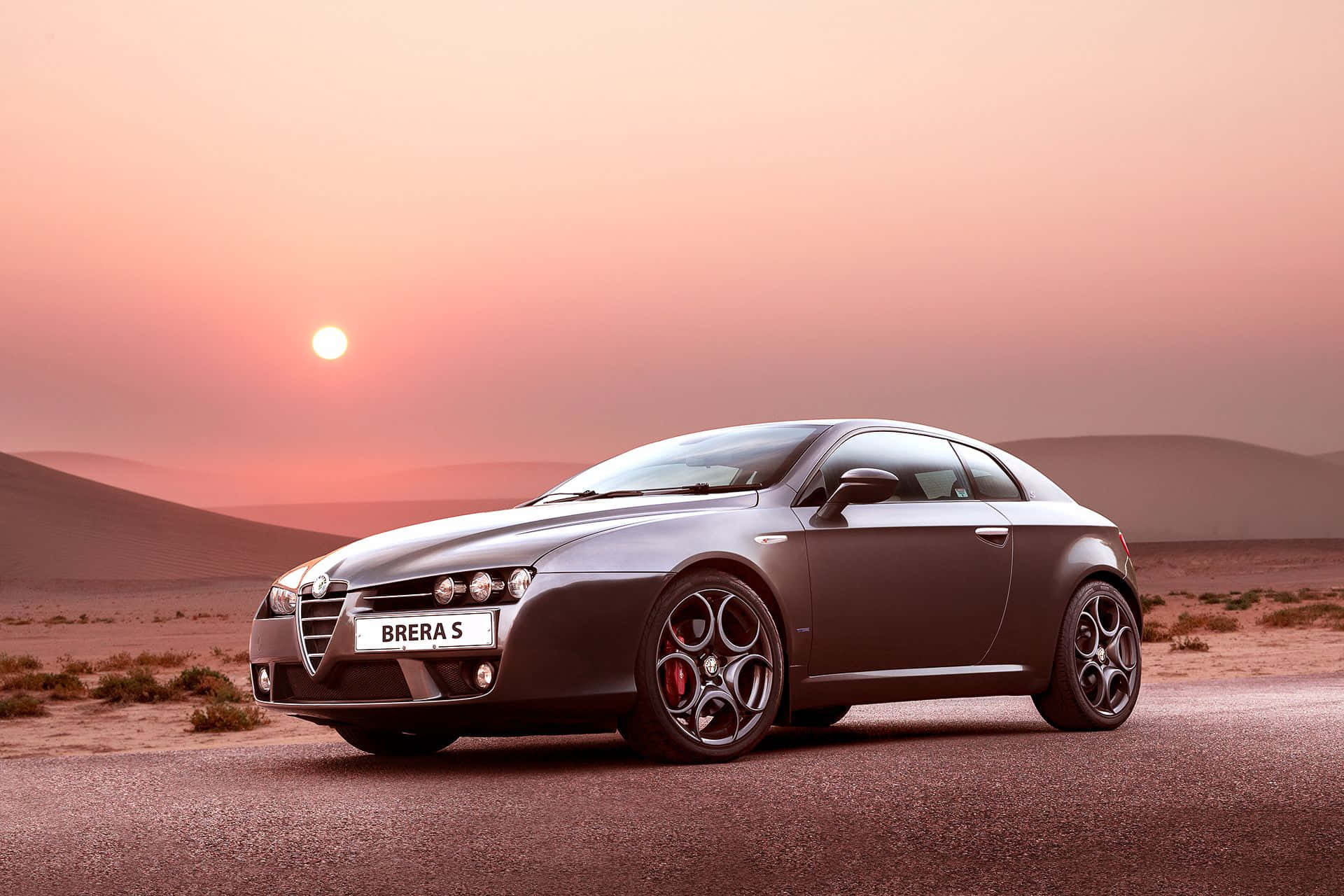 Alfa Romeo Brera 桌布 壁紙 — 一架時尚的灰色 Alfa Romeo Brera S 跑車停泊在沙漠公路上，夕陽西下，橙色的太陽在連綿沙丘後方閃耀。