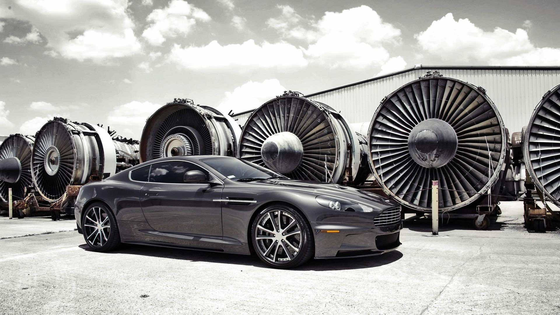 黑白 桌布 壁紙 — 一架時尚的灰色 Aston Martin 跑車停泊在巨大噴射引擎前，天空多雲。