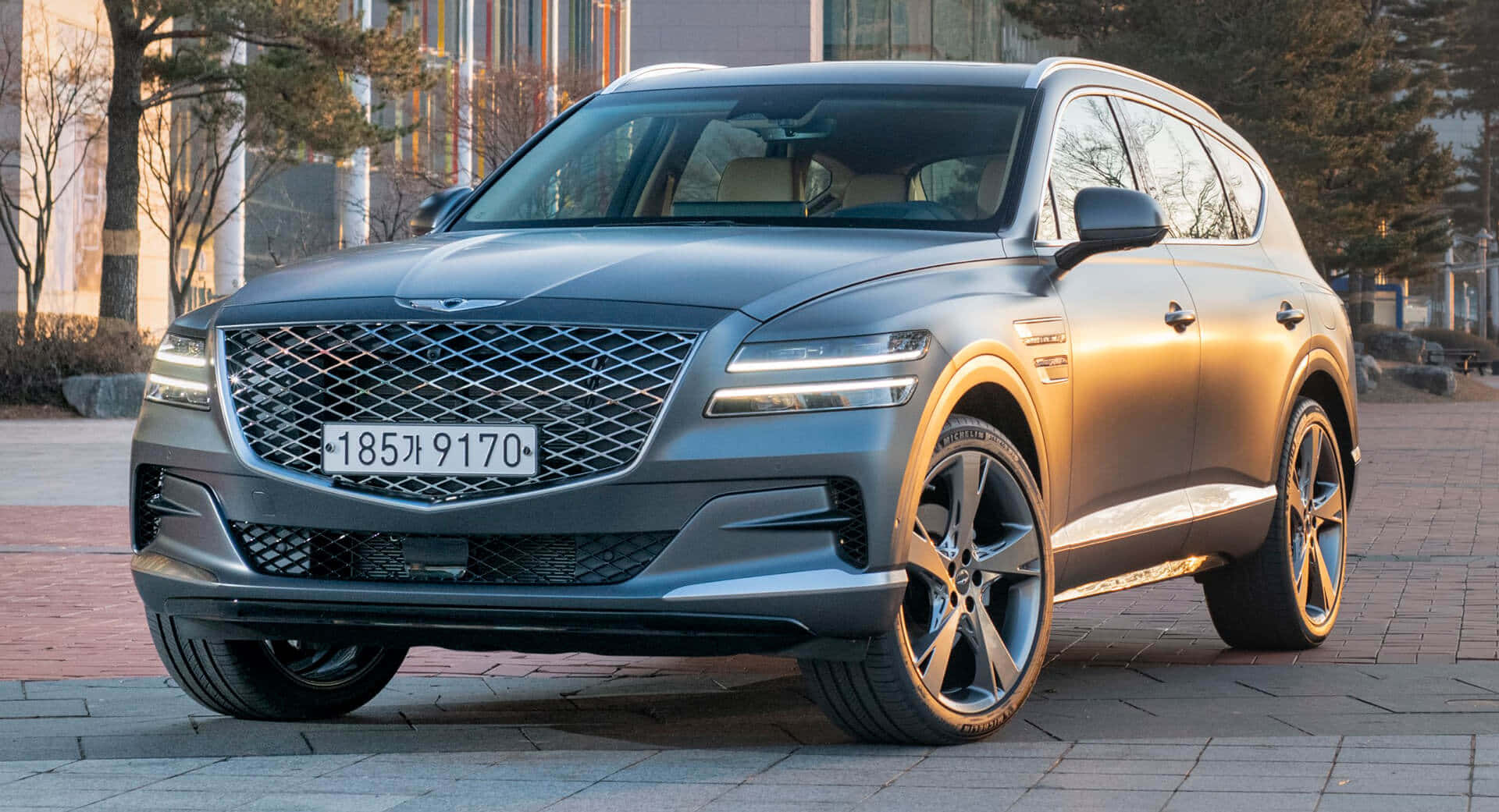 Genesis Gv80 桌布 壁紙 — 一架時尚的灰色 Genesis GV80 SUV 停泊在鋪砌的廣場上，周圍有樹木和建築物，沐浴在柔和的晨光中。