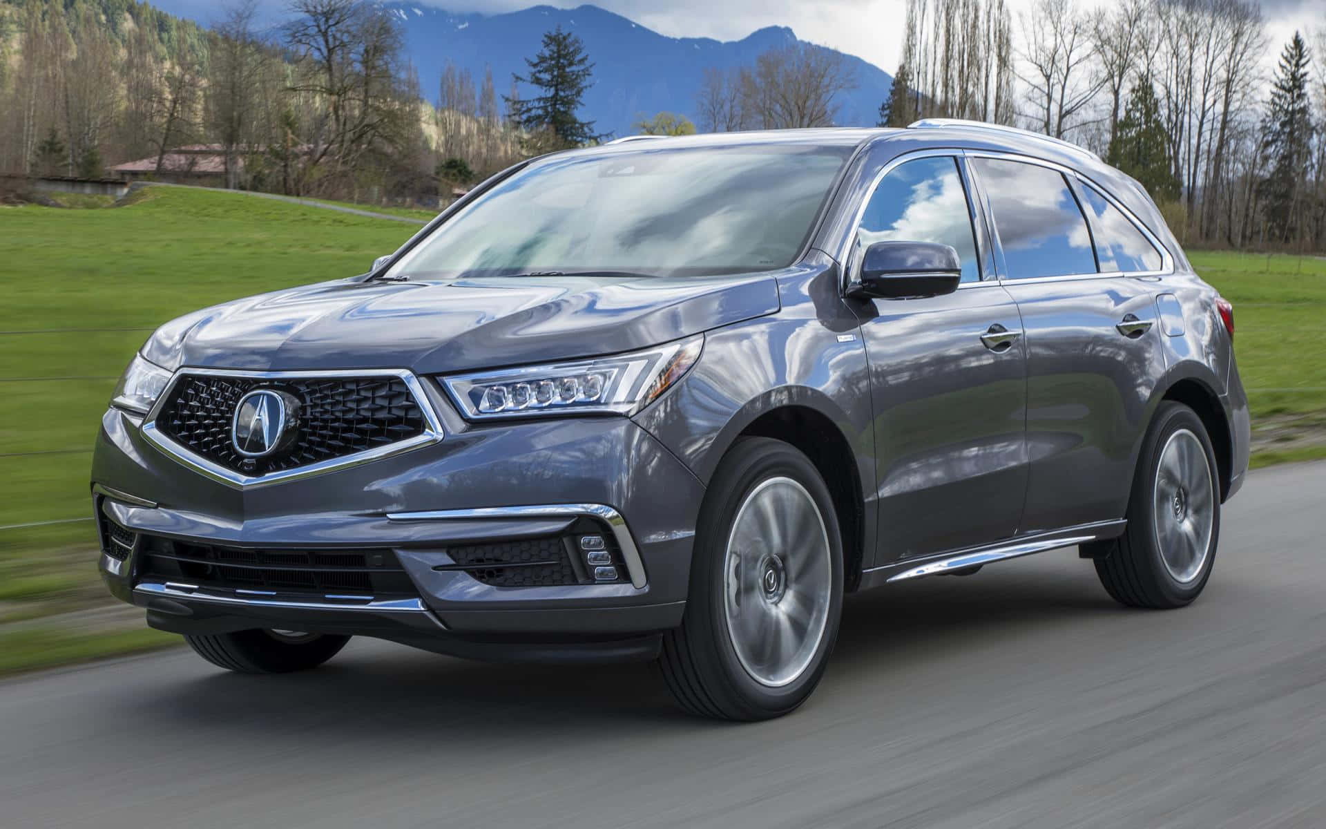 Acura Mdx 桌布 壁紙 — 一架時尚的灰色Acura SUV沿著蜿蜒的道路行駛，兩旁是茂密的綠色山丘和遠處的山脈，天空多雲。