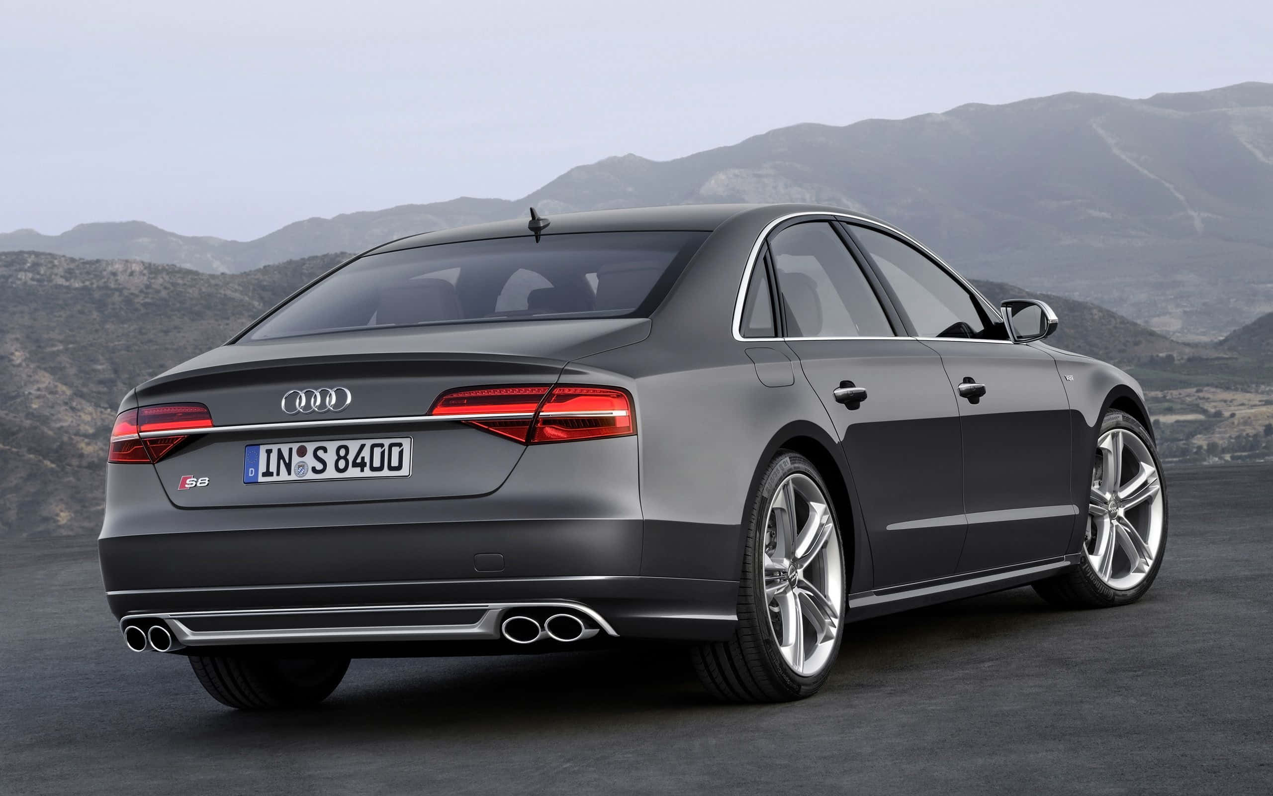 Audi S8 桌布 壁紙 — 一架時尚的灰色Audi S8房車停泊在鋪砌道路上，背景是崎嶇的山脈，天空朦朧。
