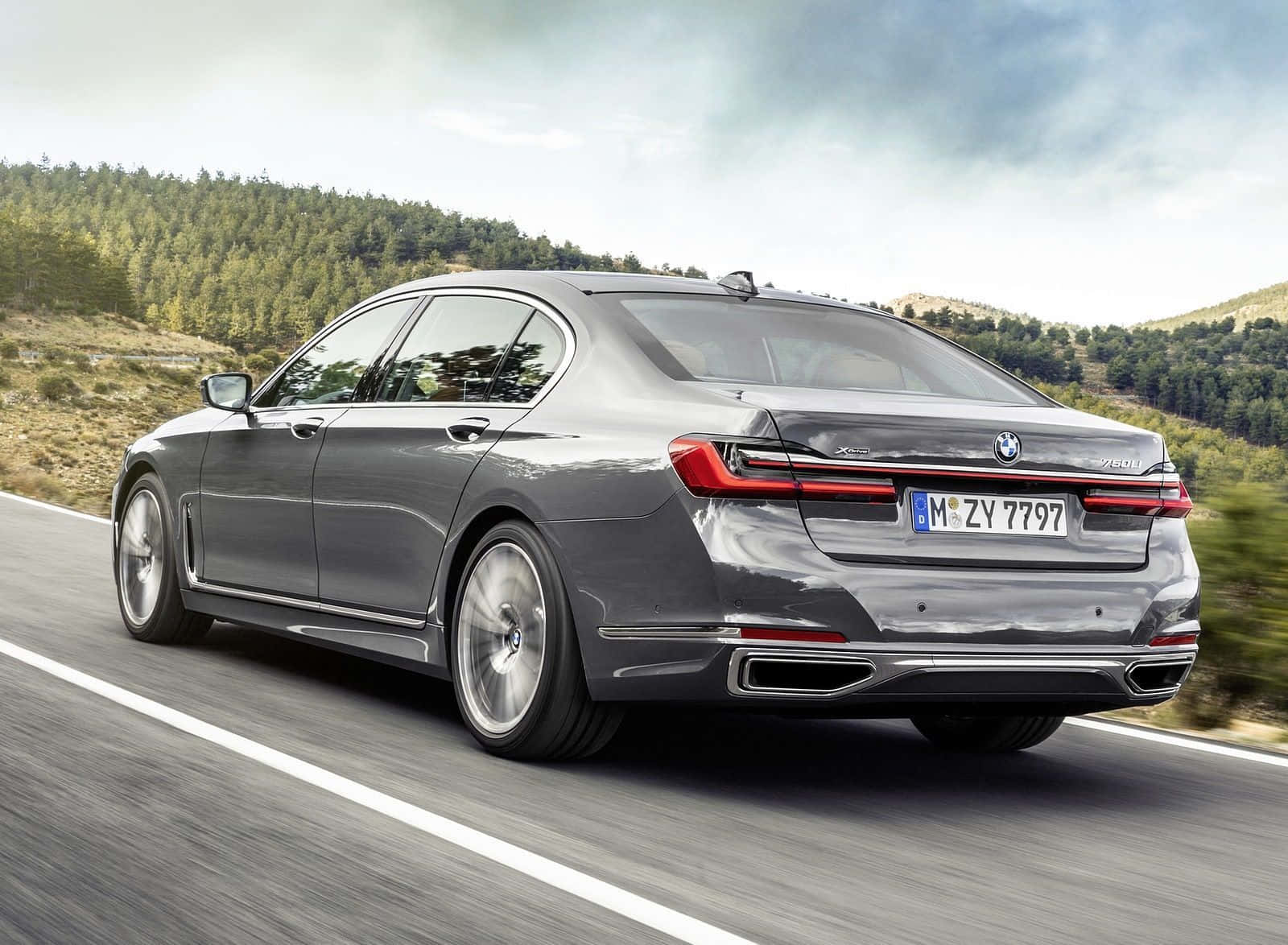 Bmw 7 Series 桌布 壁紙 — 一架時尚的灰色BMW 7系轎車沿著蜿蜒的山路行駛，兩旁是森林覆蓋的山丘，天空多雲。