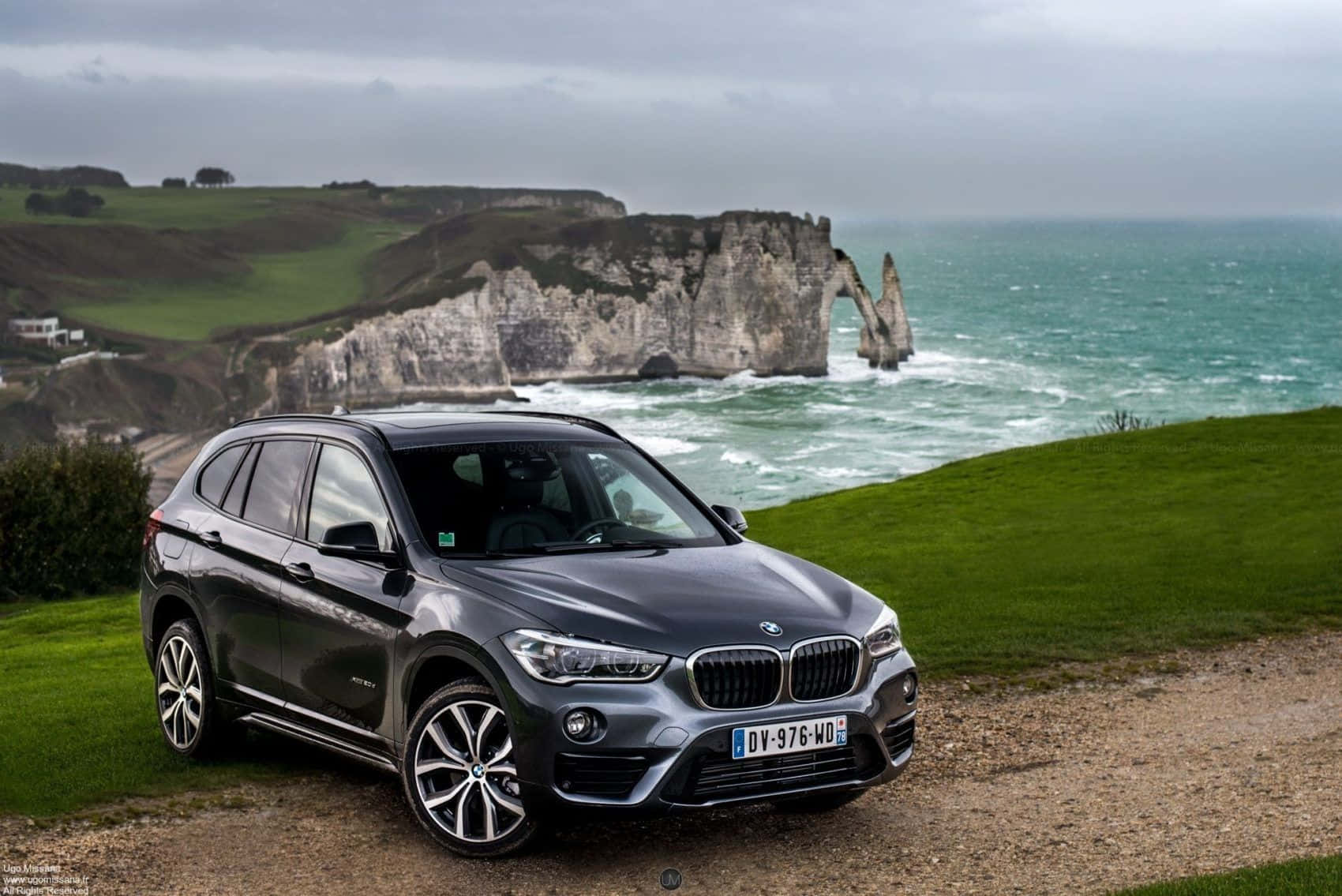 Bmw X1 桌布 壁紙 — 一架時尚的灰色BMW SUV停泊在俯瞰海洋的草地上懸崖上，背景是標誌性的白色白堊懸崖和拱門，天空多雲。