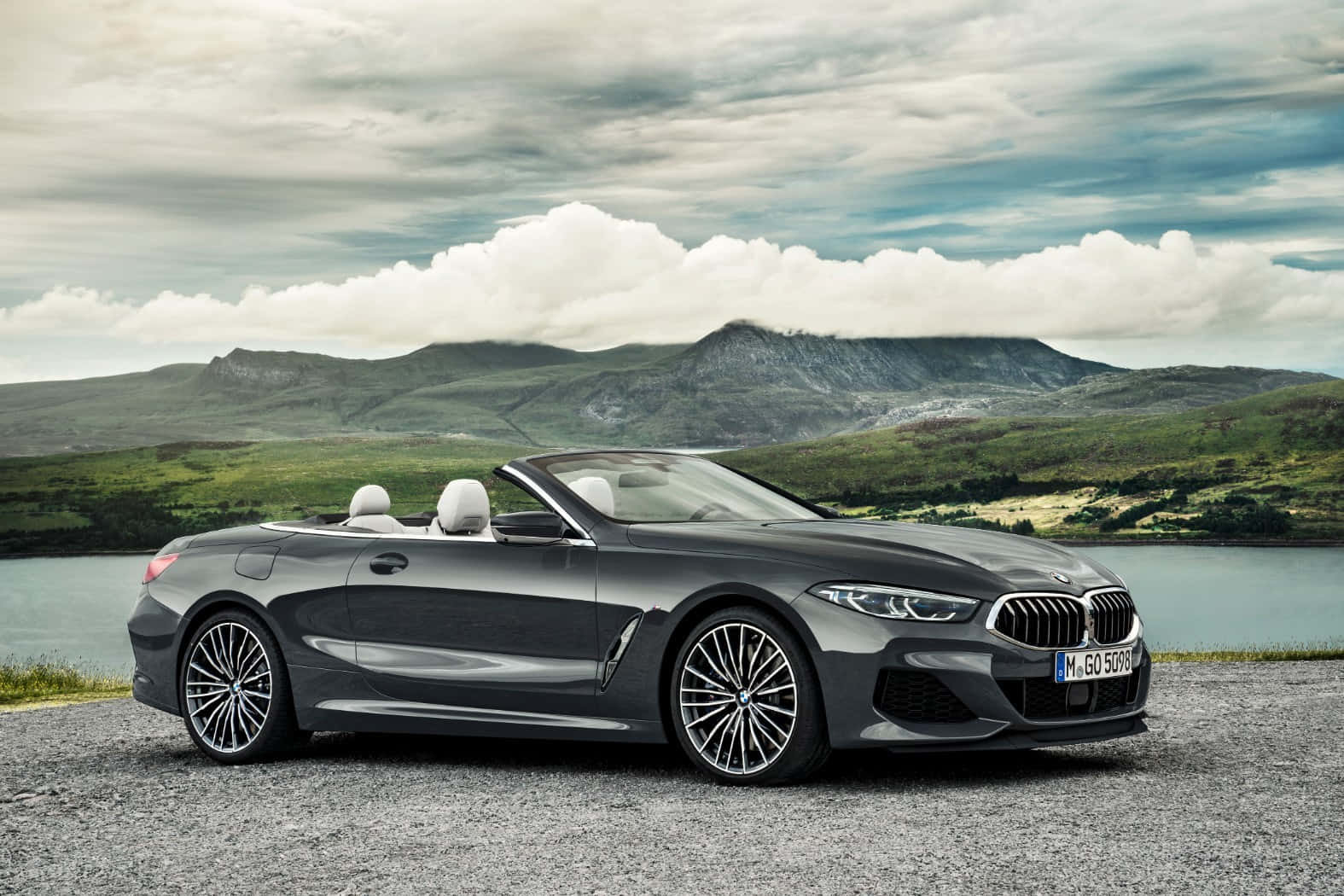 Bmw 8 Series 桌布 壁紙 — 一架時尚的灰色BMW開篷車停泊在湖邊的礫石岸上，背景是起伏的綠色山丘和多雲的天空。