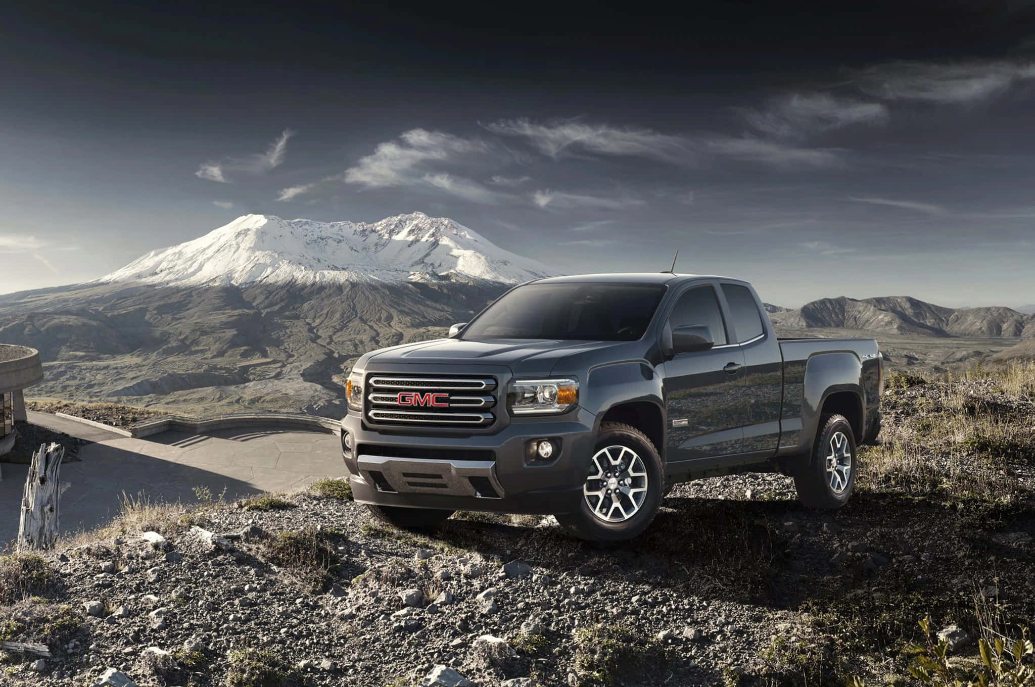 Gmc Canyon 桌布 壁紙 — 一架時尚的灰色GMC Sierra皮卡停泊在岩石觀景點上，背後有白雪覆蓋的山峰在戲劇性的多雲天空下升起。