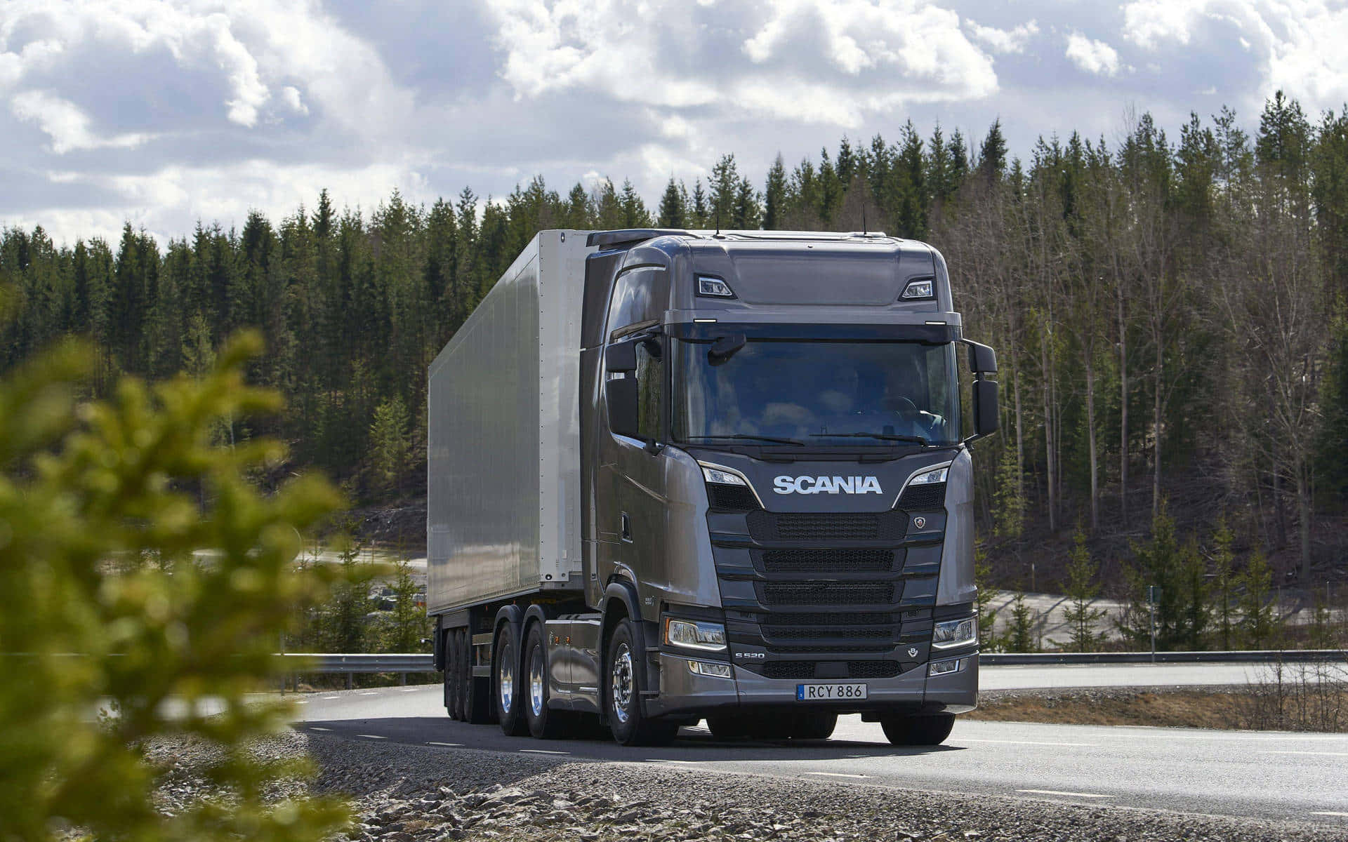 貨車 桌布 壁紙 — 一架時尚的灰色Scania半掛卡車拖著銀色拖車，在部分多雲的天空下行駛在穿過森林景觀的瀝青路上。