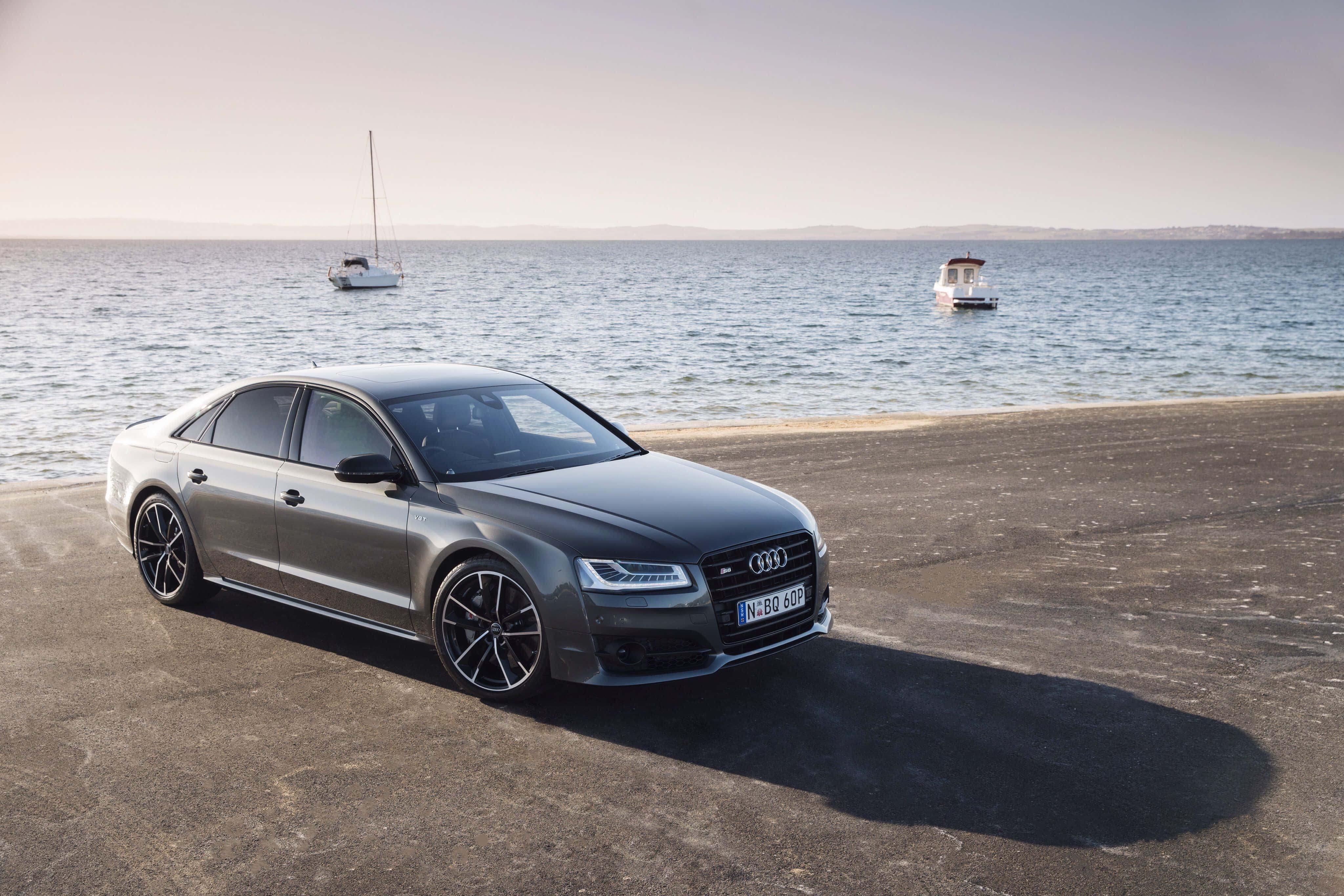 Audi S8 桌布 壁紙 — 一架時尚的灰色奧迪房車停泊在陽光普照的混凝土碼頭上，背景有兩艘船在平靜的藍色水面上漂浮。