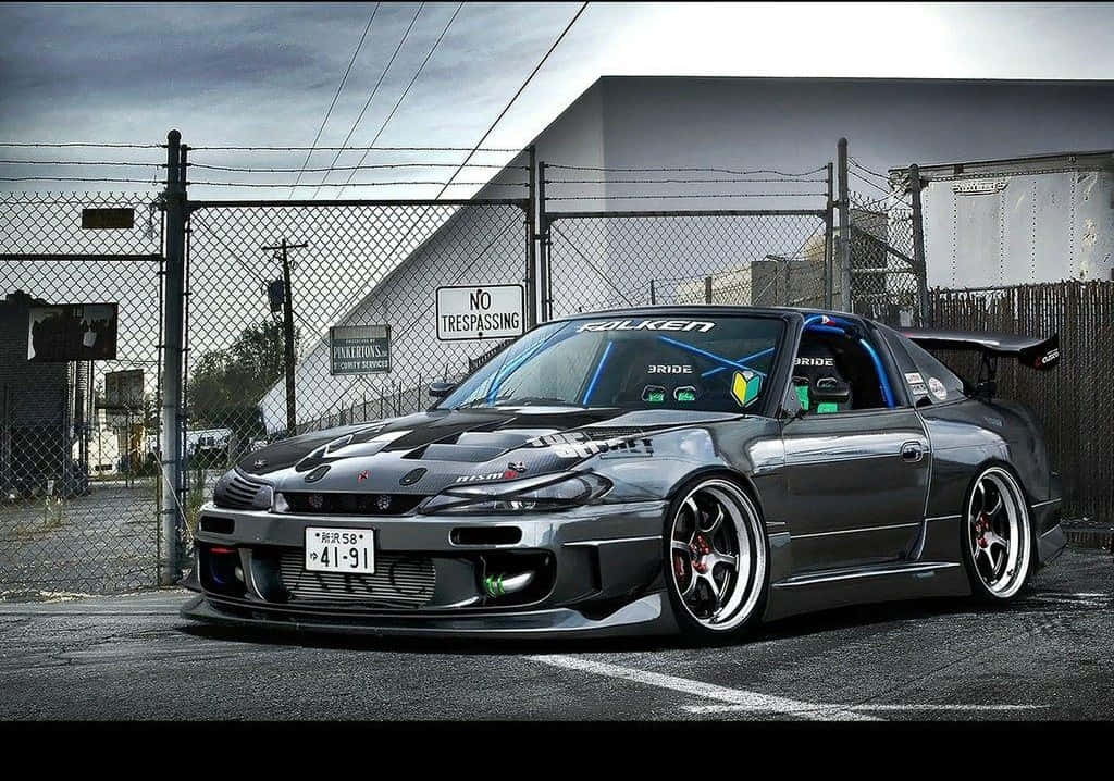 Tuned Car 桌布 壁紙 — 一架時尚的灰色日產S14漂移車，配備定制寬體套件和紅色制動卡鉗，停泊在陰天下的鐵絲網圍欄前。