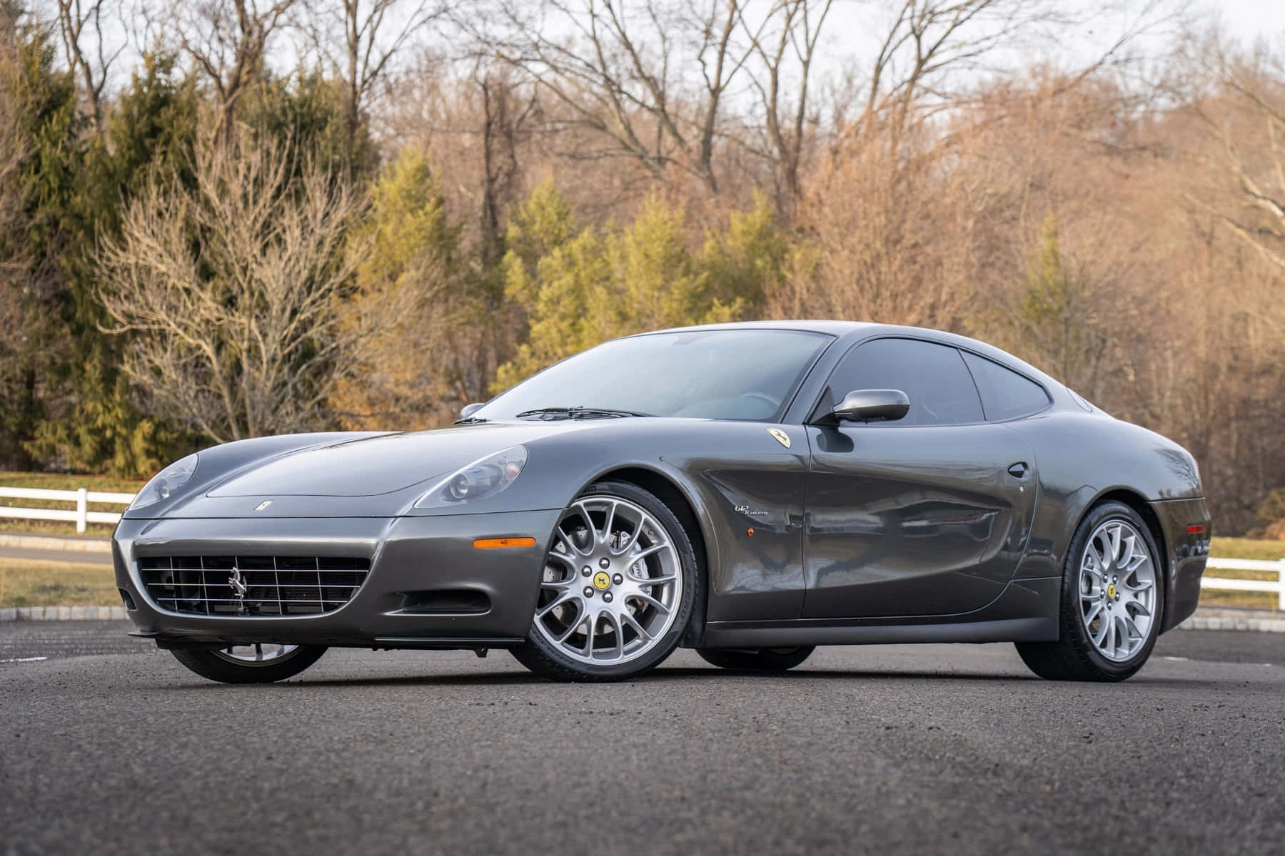 Ferrari 612 Scaglietti 桌布 壁紙 — 一架時尚的灰色法拉利612 Superamerica停在瀝青路上，背景有光禿禿的樹木和常綠植物。