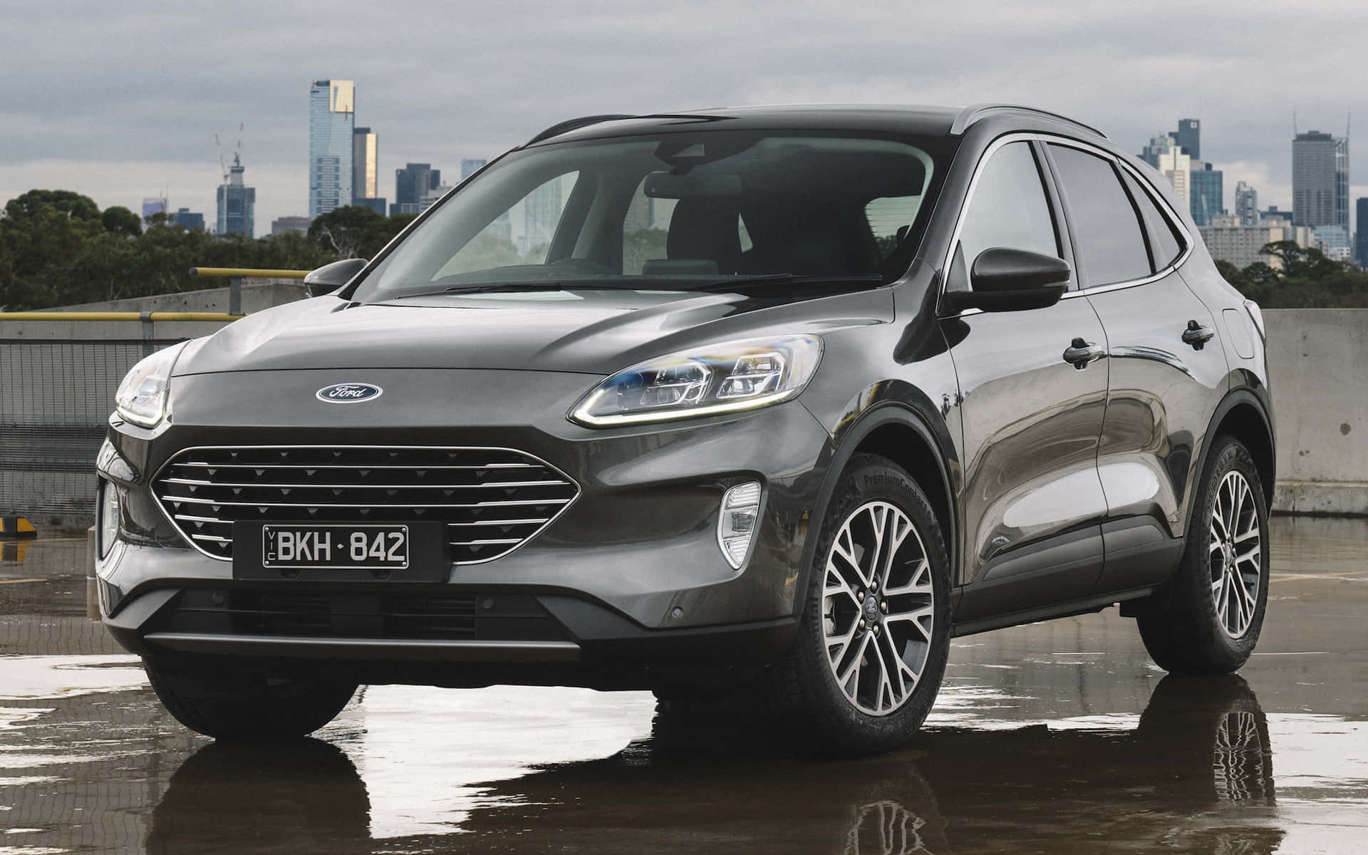 Ford Escape 桌布 壁紙 — 一架時尚的灰色福特SUV在陰天底下反射濕漉漉的路面，背景是現代化的城市天際線。