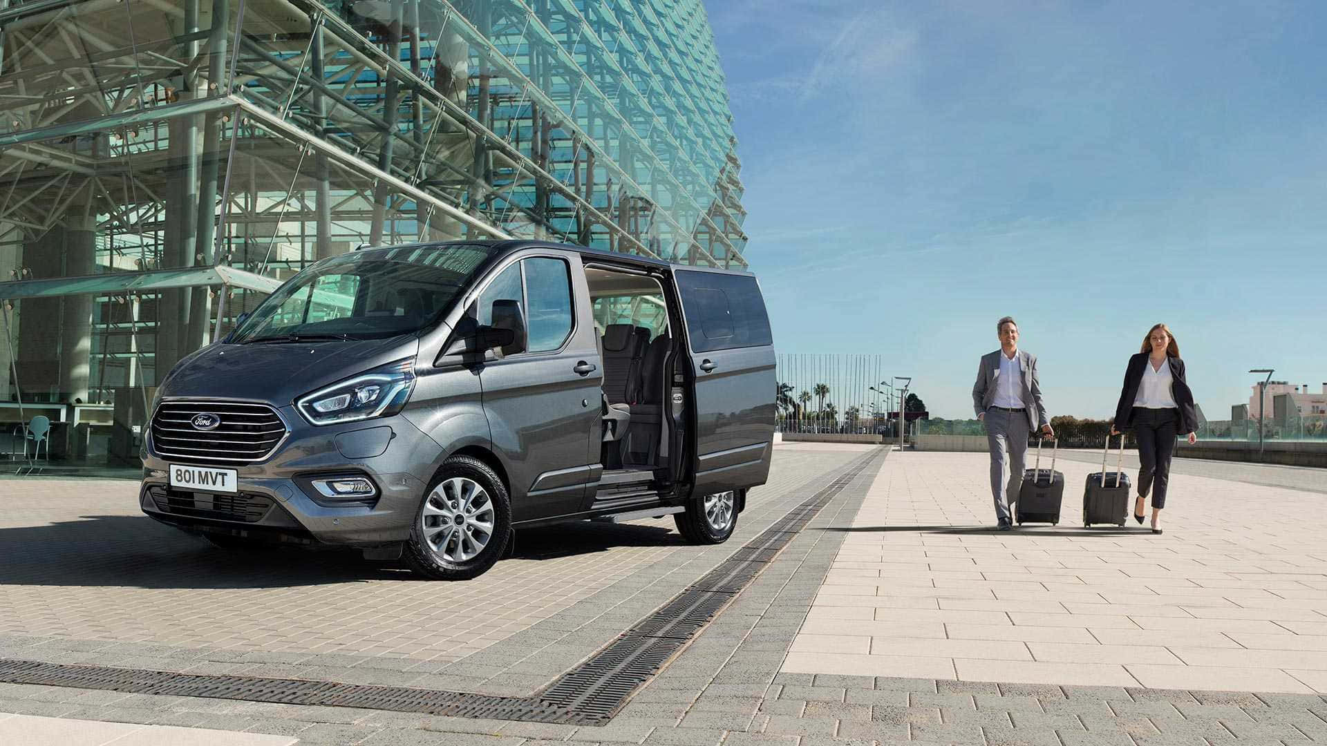 Ford Transit 桌布 壁紙 — 一架時尚的灰色福特客貨車停泊在現代玻璃建築外，兩位商務旅客在陽光明媚的日子裡拖著行李箱走過。