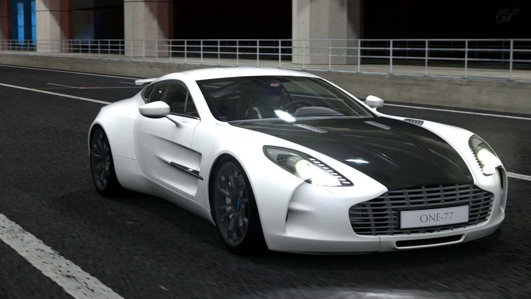Aston Martin One-77 桌布 壁紙 — 一架時尚的白色 Aston Martin One-77，帶有黑色引擎蓋條紋，在人造燈光下於黑暗賽道上疾馳。
