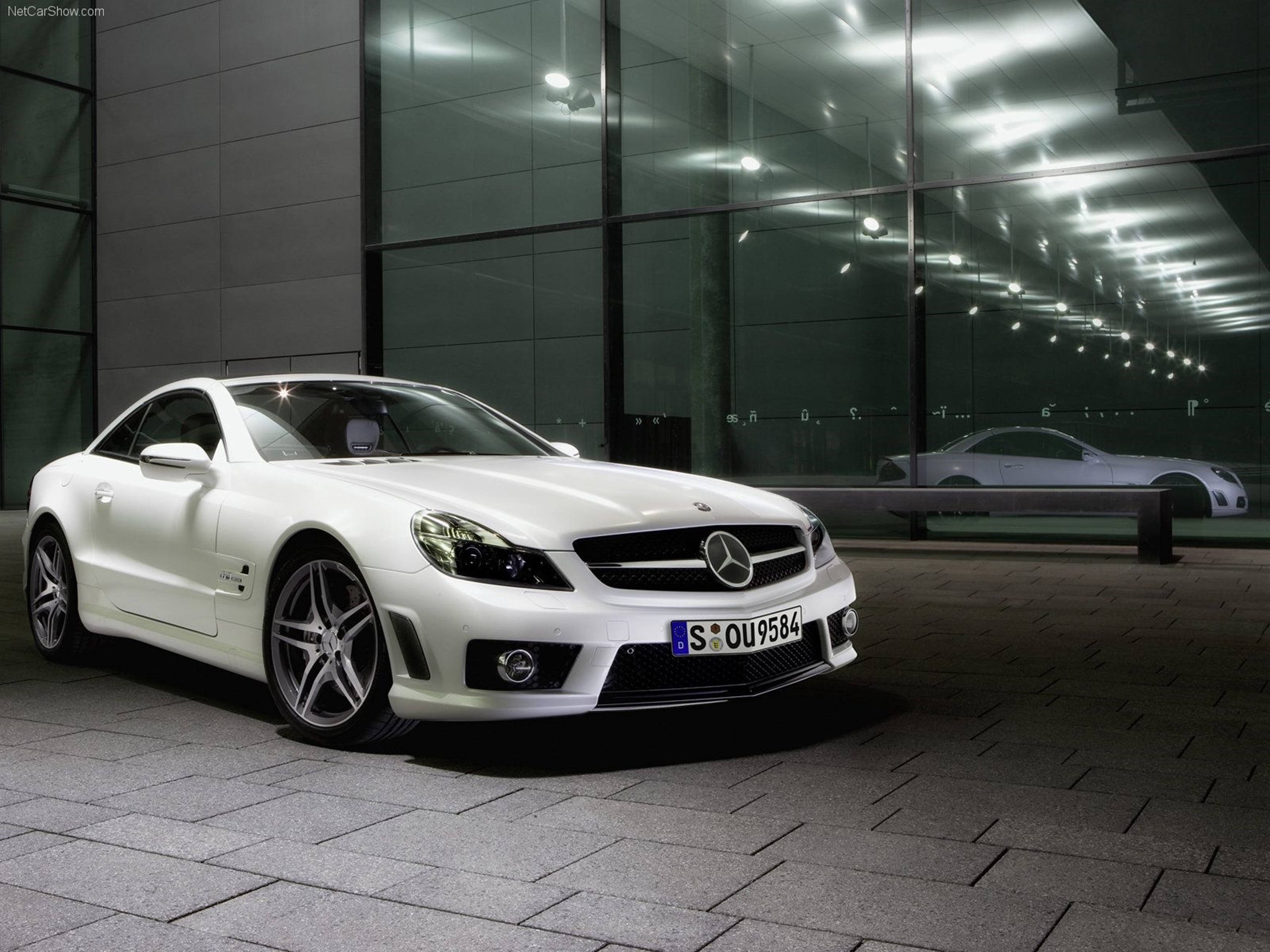 HD 桌布 壁紙 — 一架時尚的白色 Mercedes SLR McLaren 跑車在人造燈光下閃閃發光，停泊在現代玻璃建築旁邊的瓷磚廣場上。