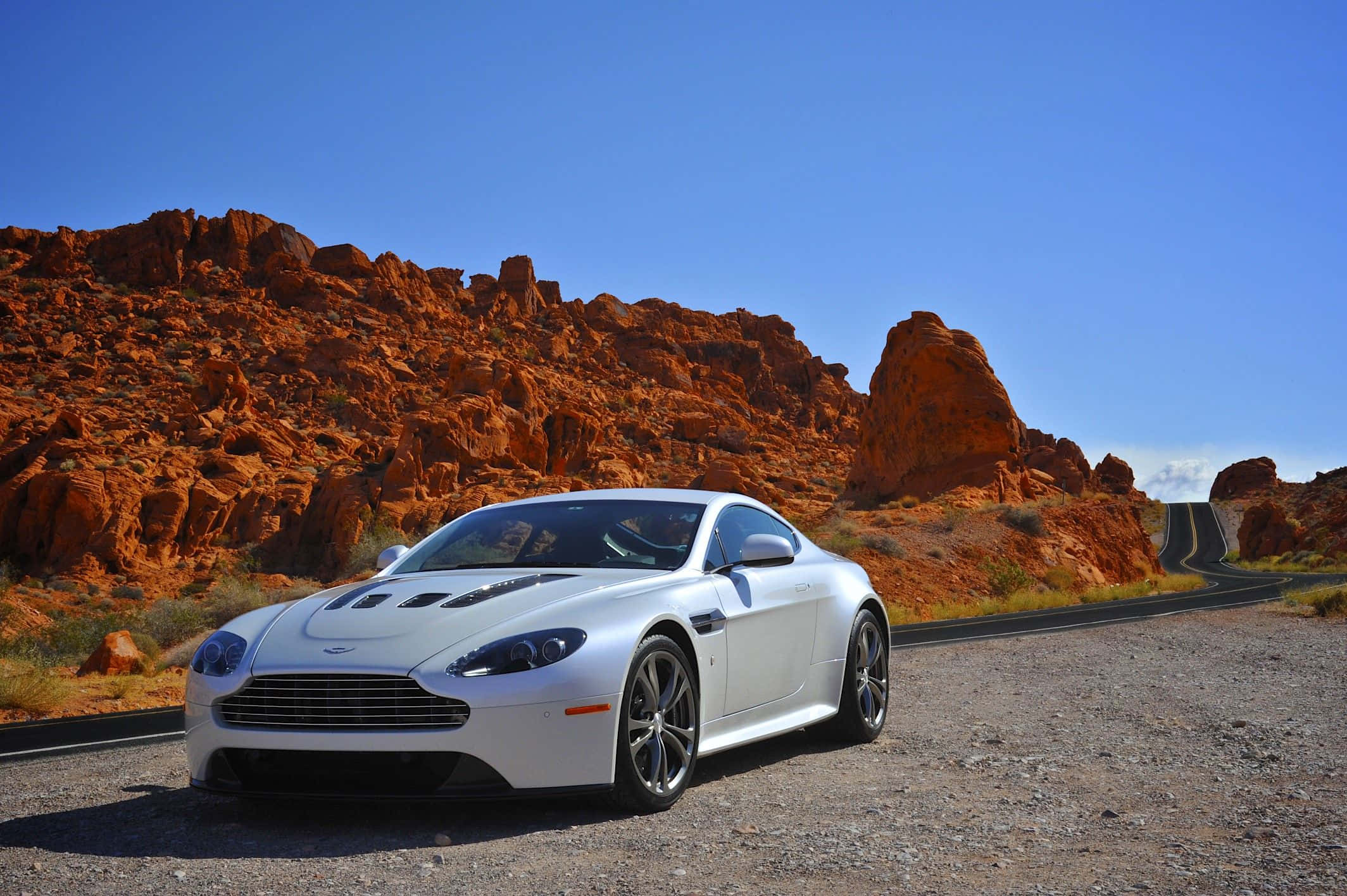 Aston Martin V12 Vantage 桌布 壁紙 — 一架時尚的白色Aston Martin跑車停泊在沙漠公路上，背景是紅色岩石地貌，天空清澈湛藍。