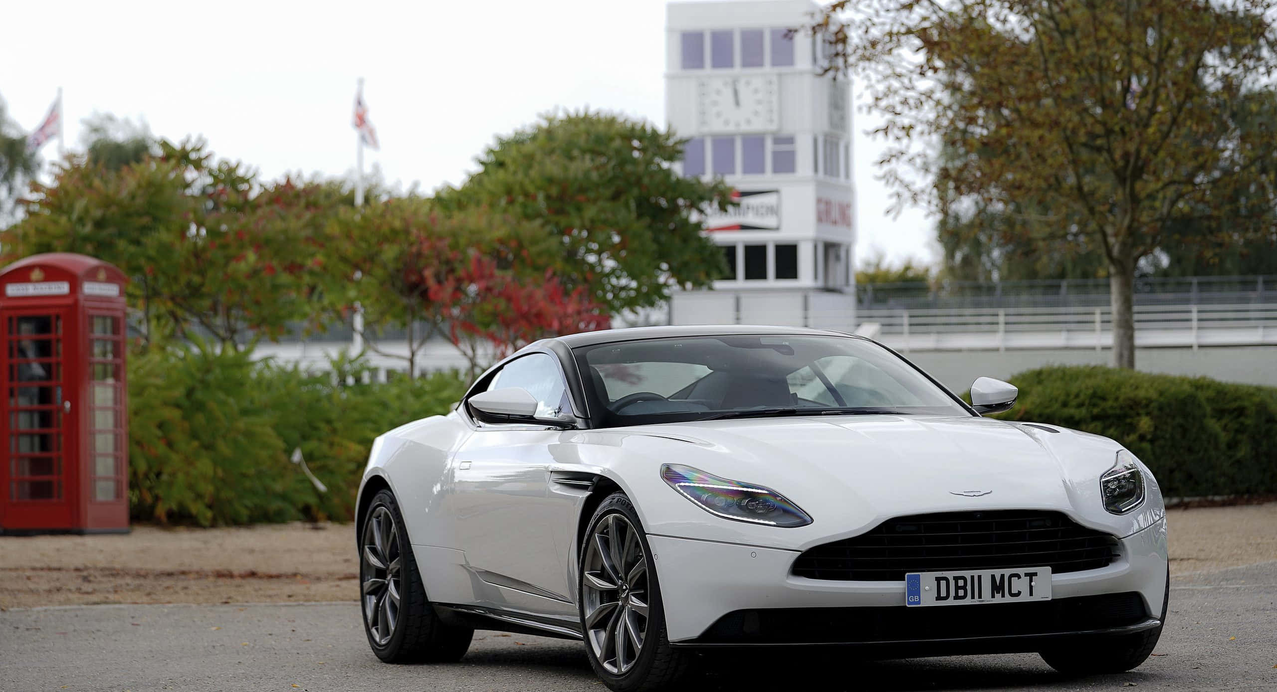 Aston Martin Db11 桌布 壁紙 — 一架時尚的白色Aston Martin DB11停泊在經典的紅色英國電話亭旁，背景有秋樹和鐘樓。