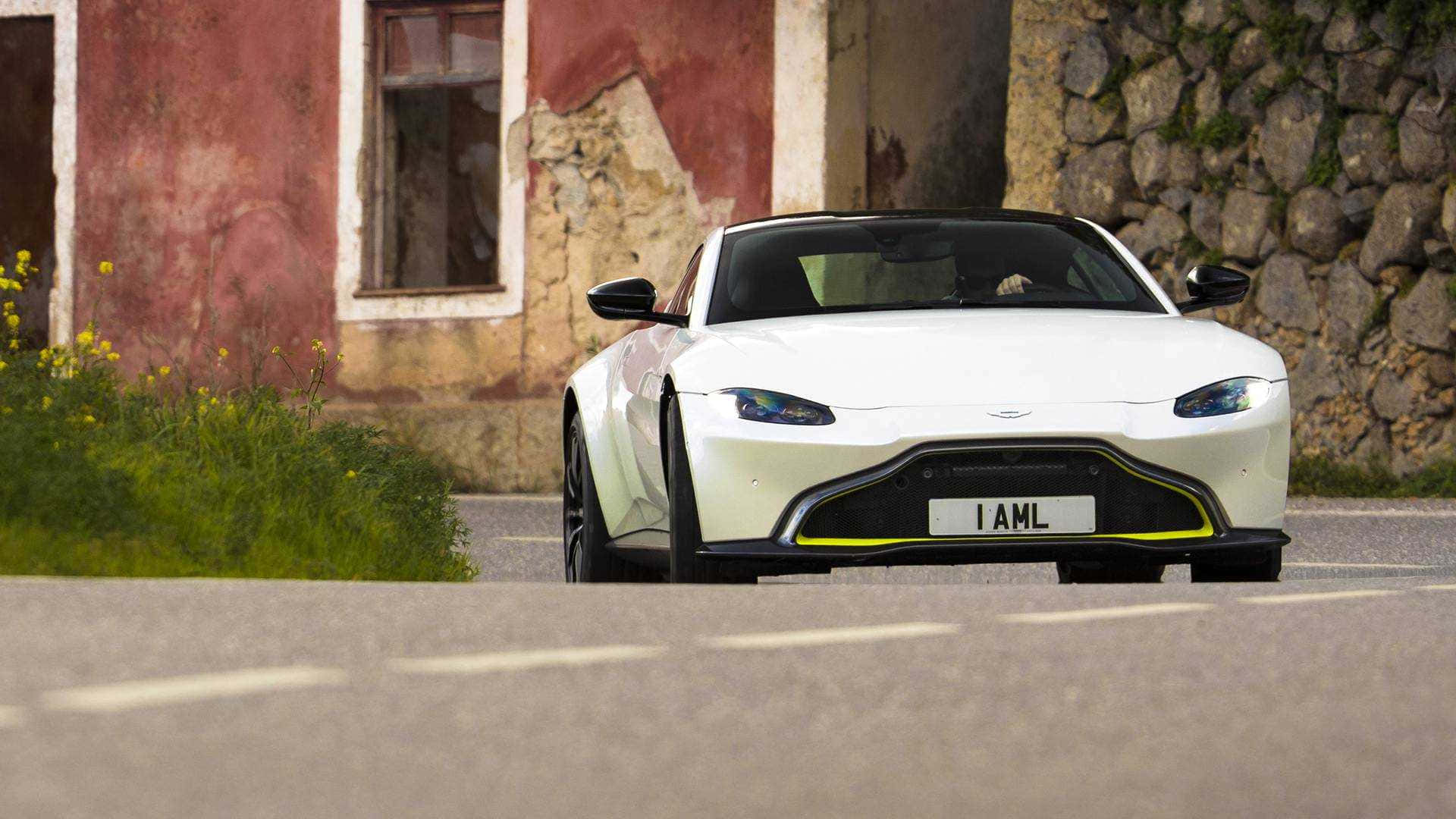 Aston Martin Vantage 桌布 壁紙 — 一架時尚的白色Aston Martin Vanquish Zagato停泊在路邊，旁邊是風化的紅色建築和石牆。