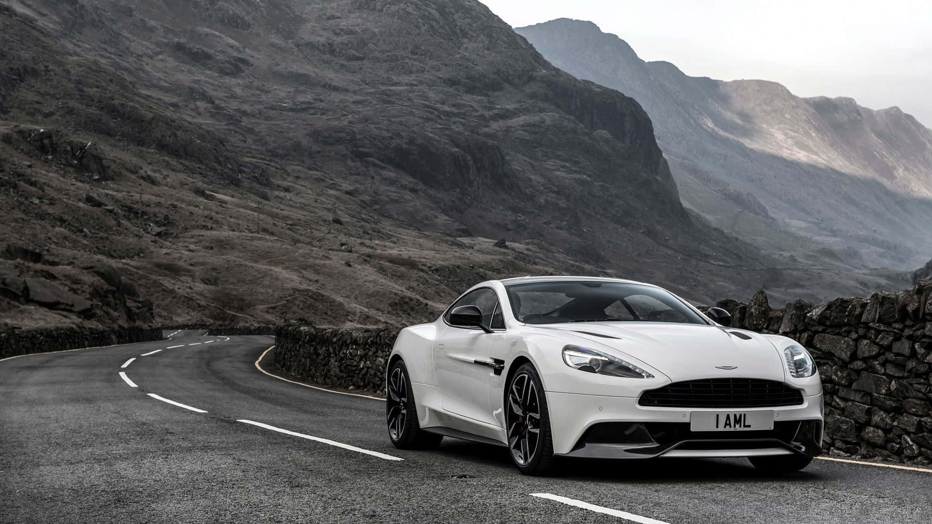 Aston Martin Vanquish 桌布 壁紙 — 一架時尚的白色Aston Martin跑車在蜿蜒的山路上行駛，兩旁是崎嶇的石牆和霧氣籠罩的山峰。