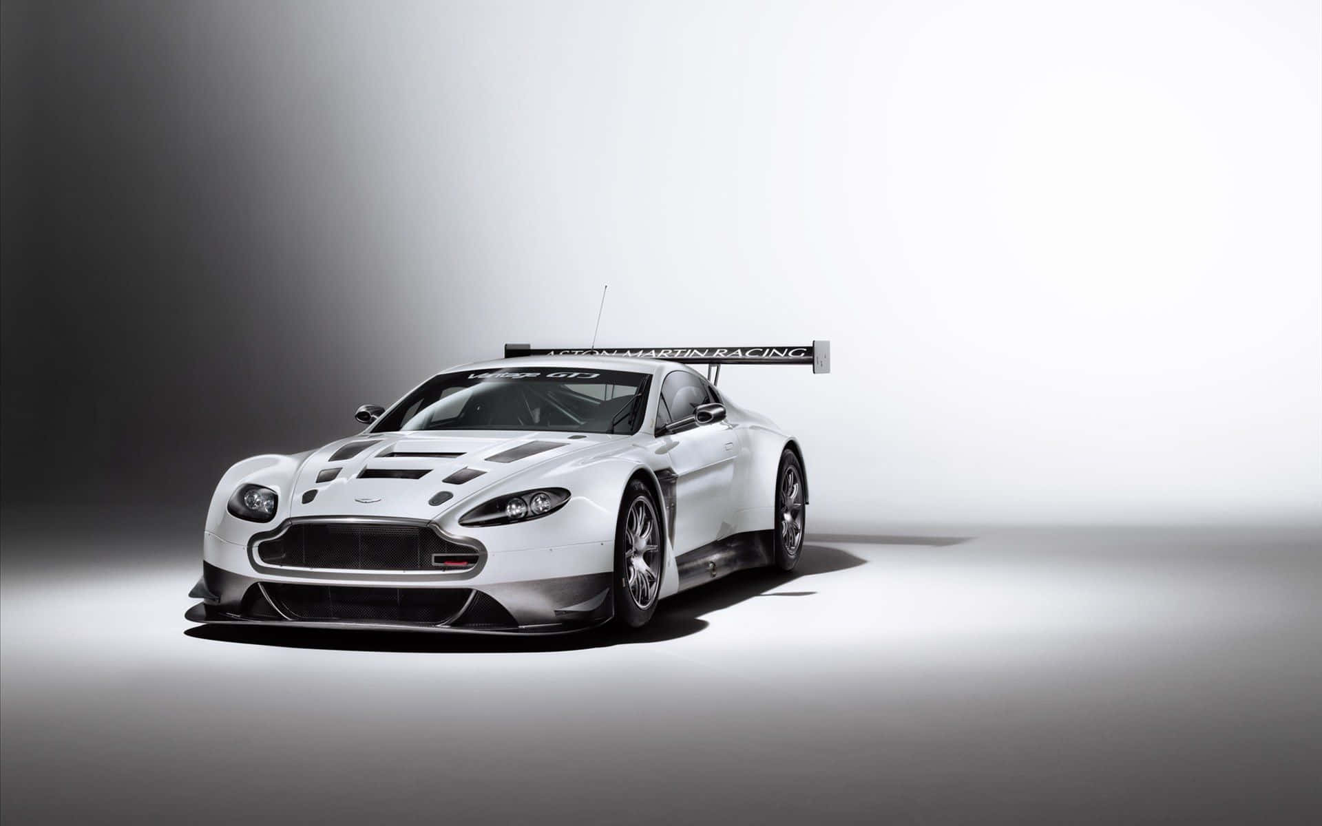 Aston Martin V12 Vantage 桌布 壁紙 — 一架時尚的白色Aston Martin賽車，帶有黑色條紋和大型後翼，在漸變灰色工作室背景上戲劇性地打光。