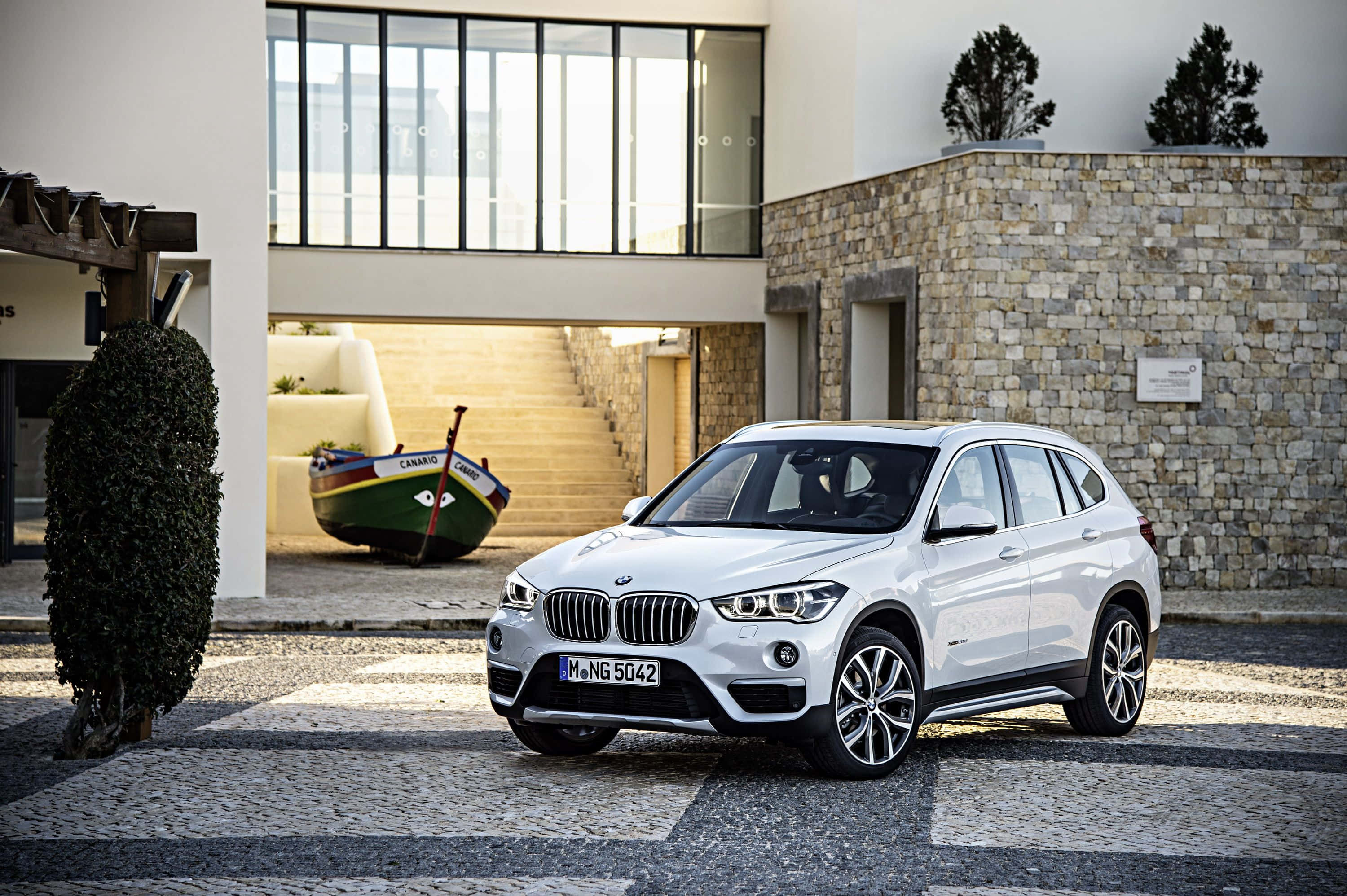 Bmw X1 桌布 壁紙 — 一架時尚的白色BMW SUV停泊在鵝卵石車道上，前面係一間現代石頭同玻璃屋，入面有裝飾船。