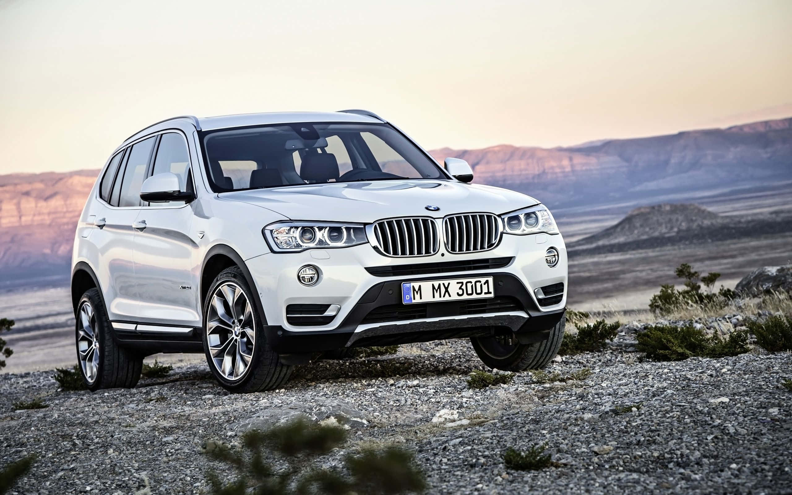 Bmw X3 桌布 壁紙 — 一架時尚的白色BMW SUV停泊在岩石地形上，背景是沙漠山脈和柔和的日落天空。