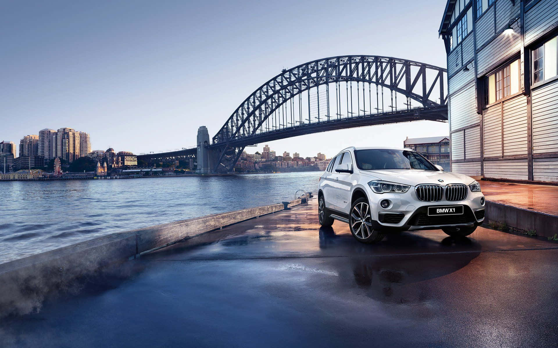 Bmw X1 桌布 壁紙 — 一架時尚的白色BMW X1 SUV停泊在濕潤的海濱碼頭，背景是標誌性的悉尼港灣大橋，天空湛藍清澈。