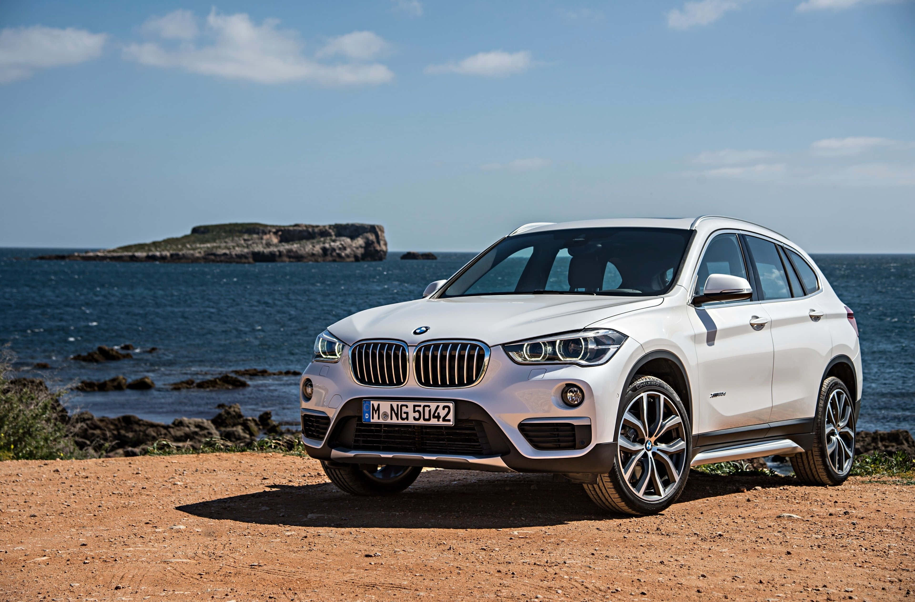 Bmw X1 桌布 壁紙 — 一架時尚的白色BMW X1 SUV停泊在陽光明媚的海岸懸崖上，俯瞰深藍色的海洋，遠處有岩石小島。